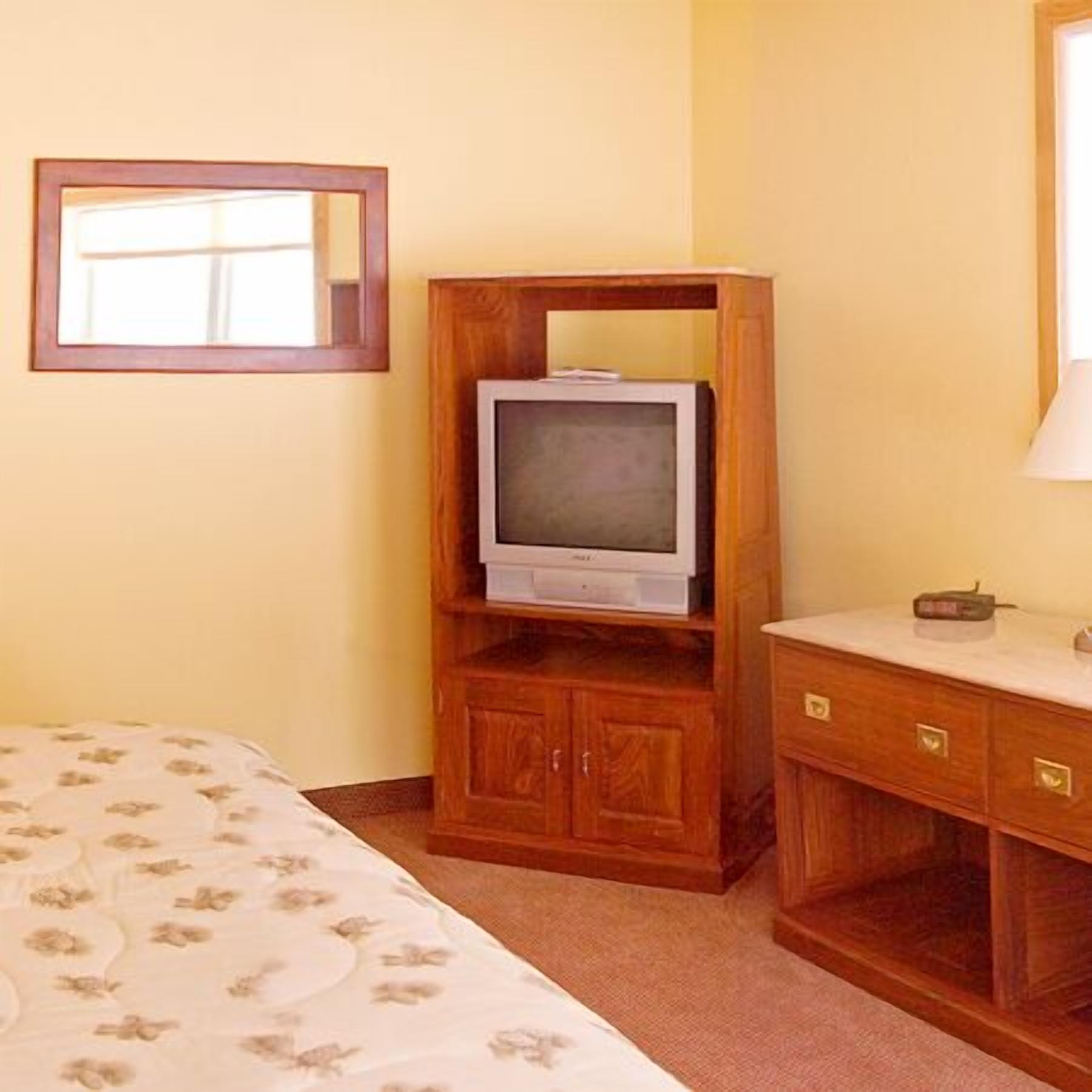 Una habitación de hotel con un mueble bajo de televisión de madera, una cama con colcha estampada y una mesita de noche con lámpara.
