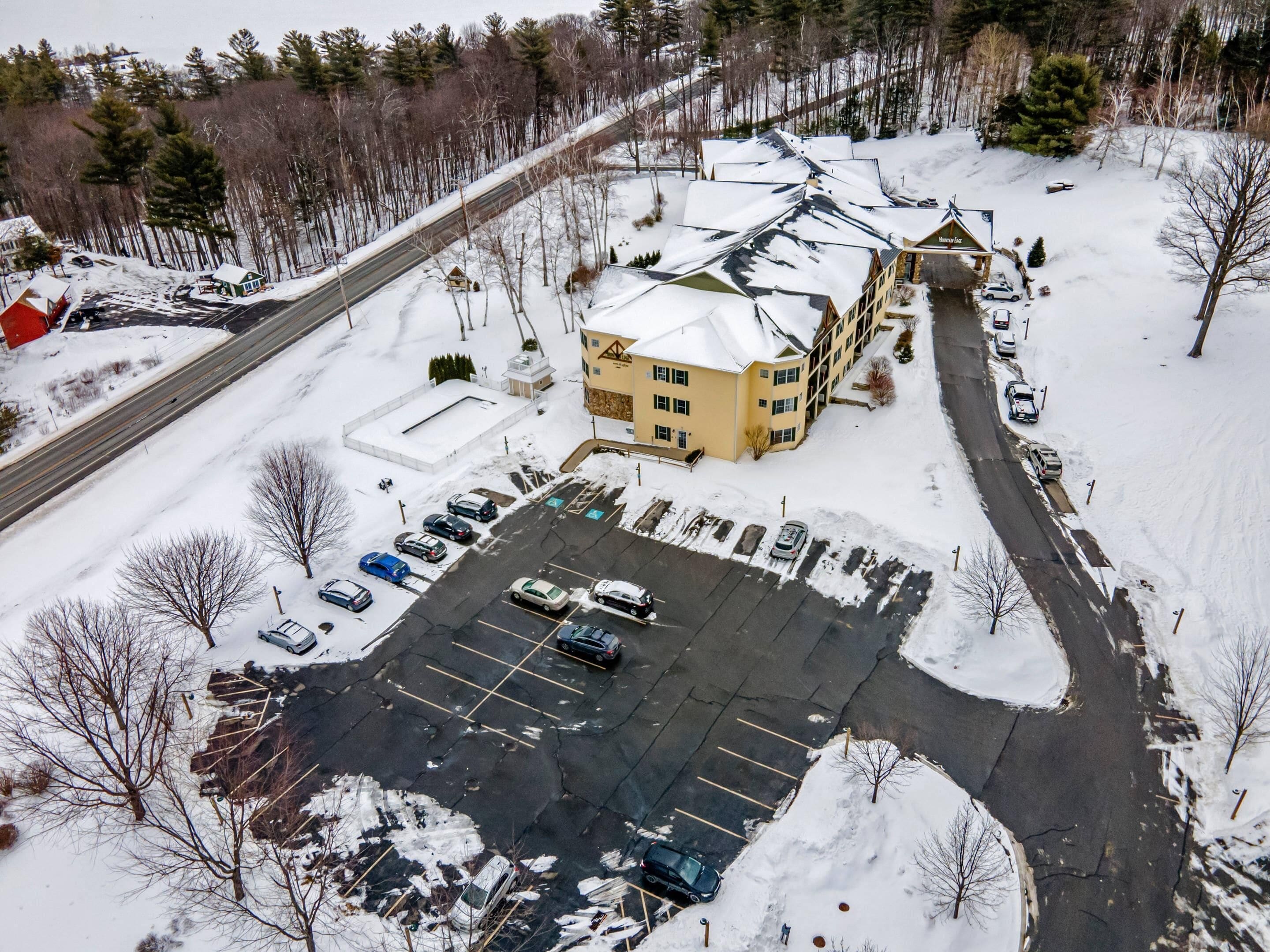 Un edificio grande con techo nevado, un estacionamiento con varios autos y una carretera junto a un área boscosa.