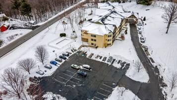 Un edificio grande con techo nevado, un estacionamiento con varios autos y una carretera junto a un área boscosa.