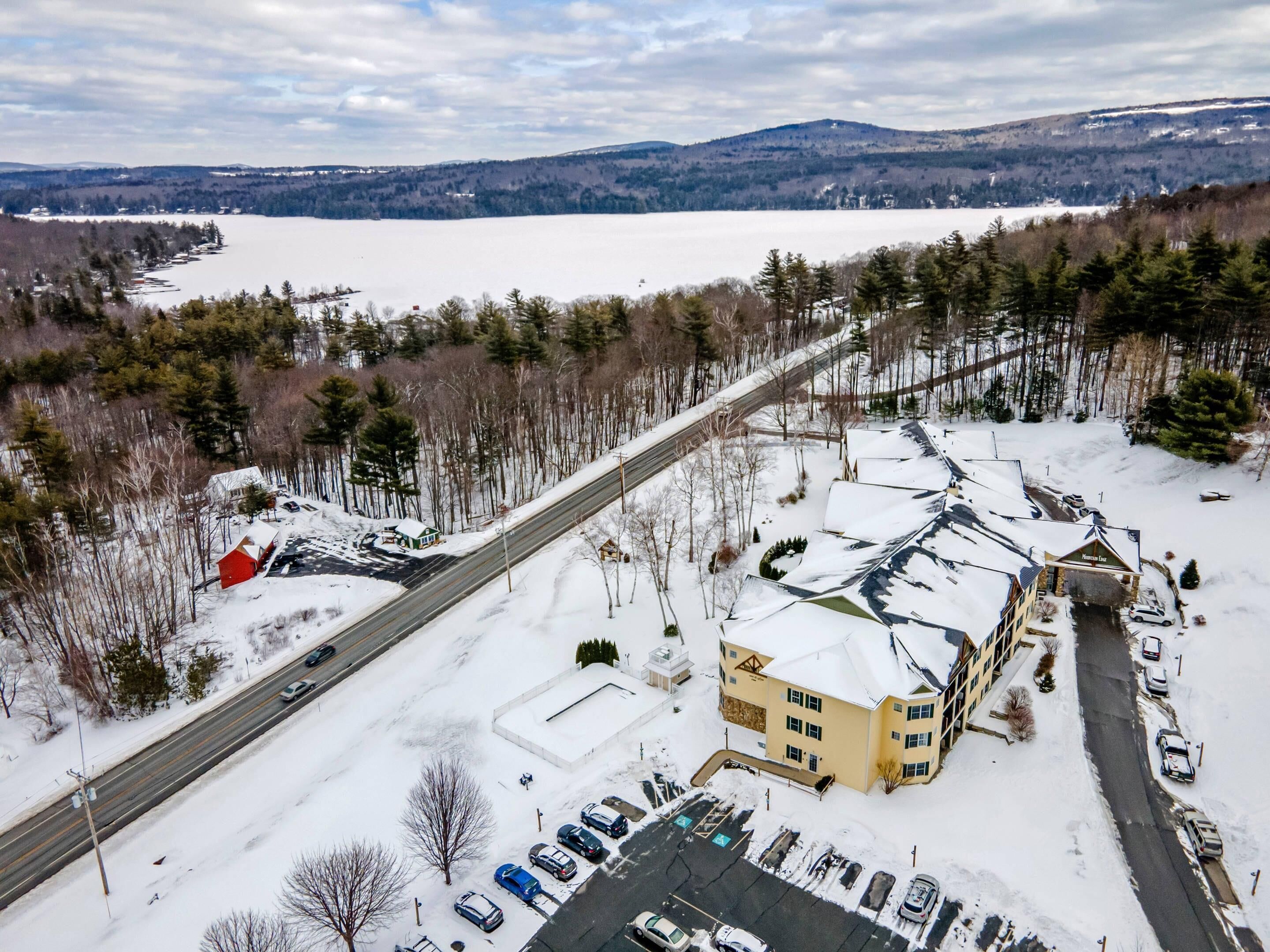 En snedækket parkeringsplads med flere biler, en bygning med skiltet "Mountain Lodge" og en vej, der løber langs siden.