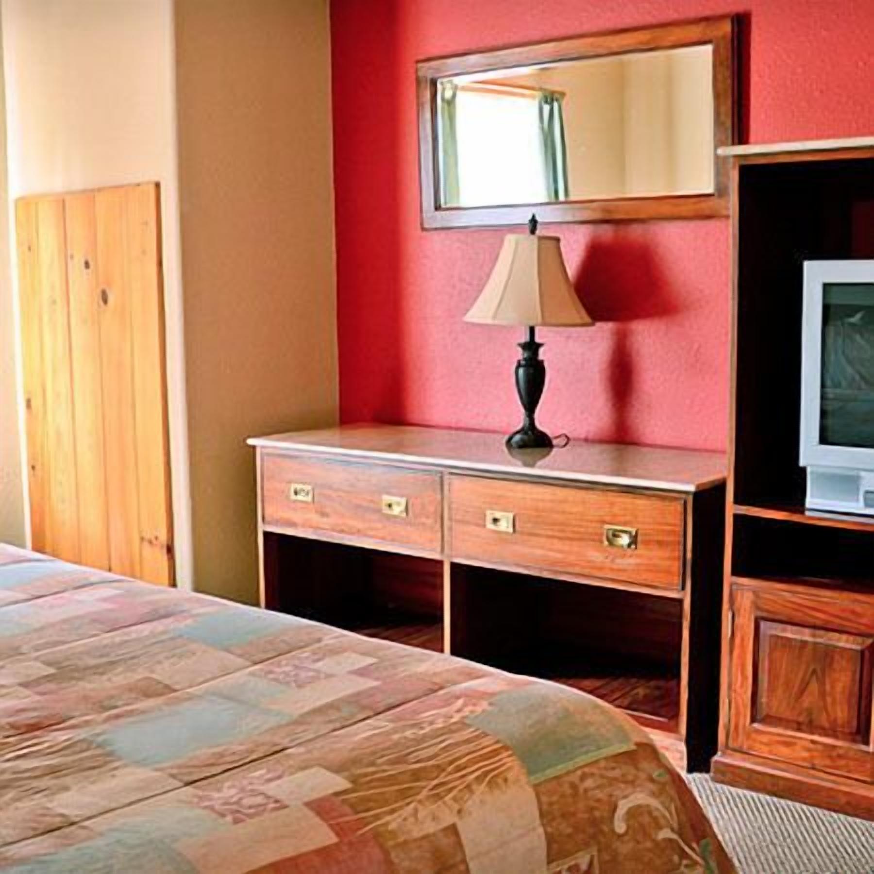 A bedroom with a bed, a wooden dresser, a lamp, a television, and a mirror.