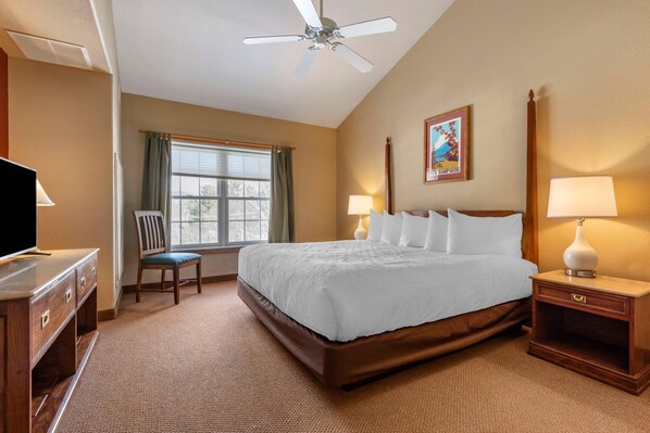 A hotel room with a bed, two bedside tables with lamps, a chair, a desk with a television, a ceiling fan, and a window with curtains. - Mountain Edge Suites at Sunapee, an Ascend Collection Hotel (Newbury)