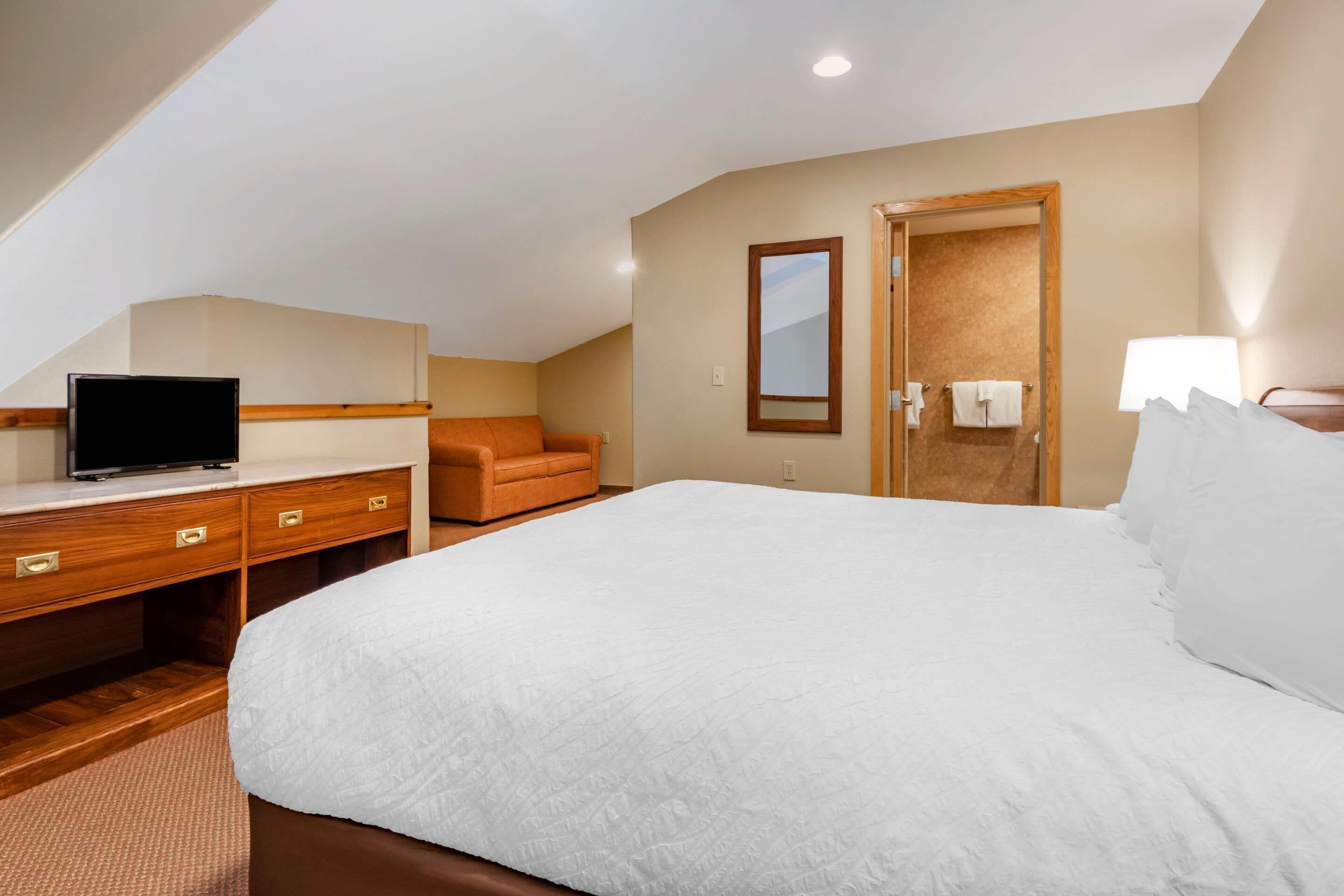 A hotel room with a large bed, a desk with a television, a chair, a door with a towel hanging, and a mirror on the wall.