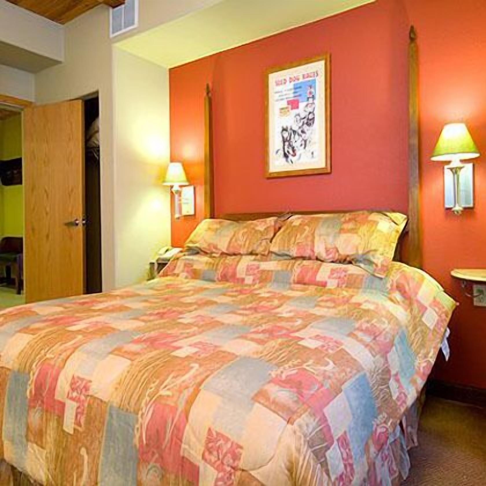 A bedroom with a bed, two bedside lamps, a framed poster on the wall, and a door to another room.