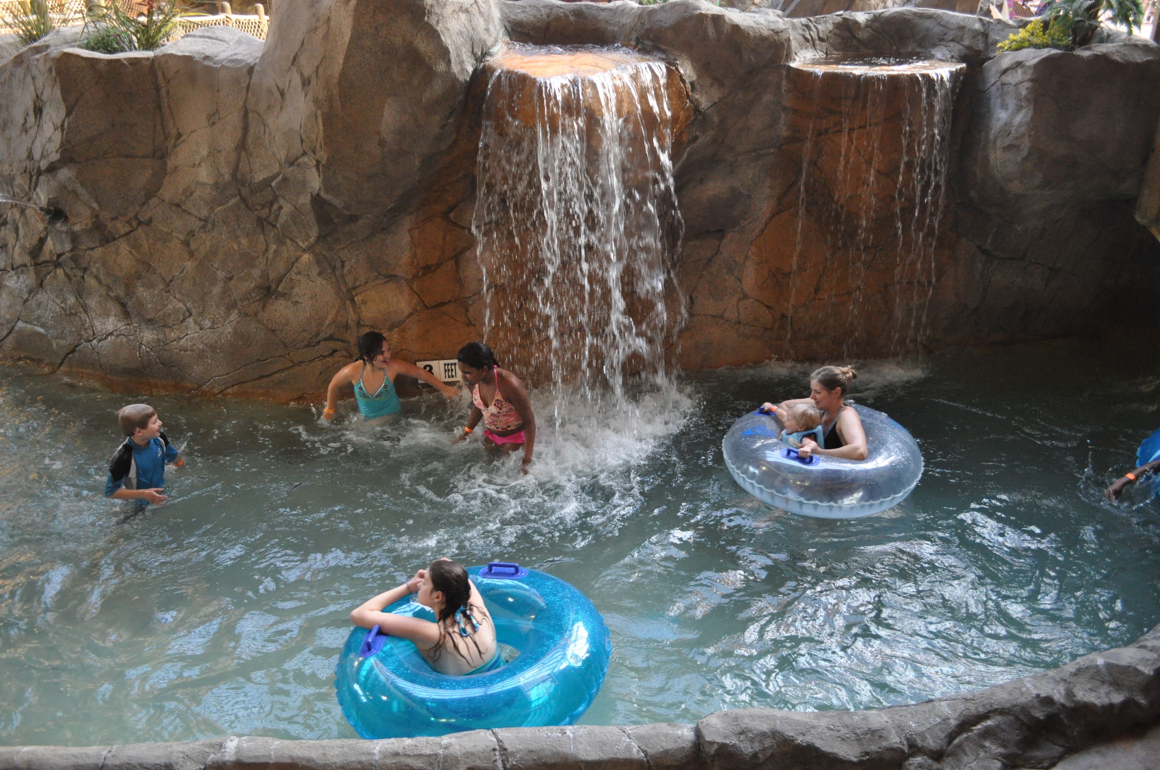 Um parque aquático com cachoeira, boias infláveis e pessoas se divertindo na água.