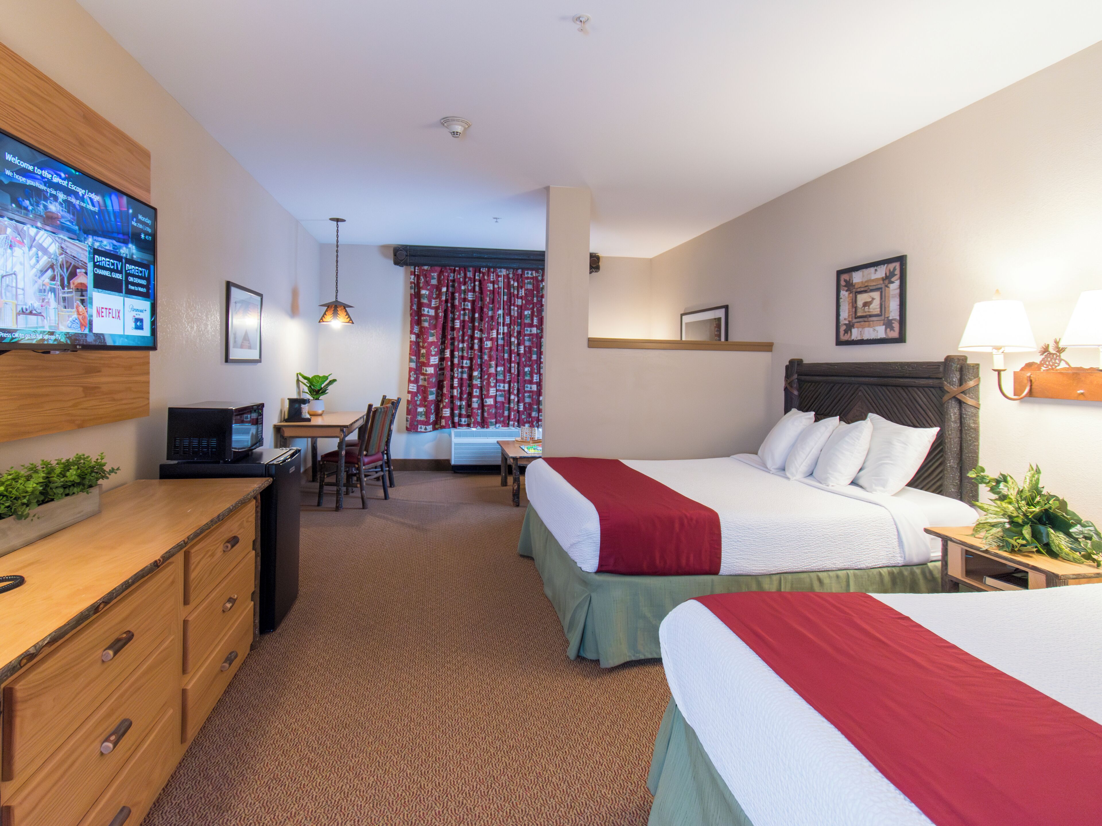 A hotel room with two beds, a desk with a computer, a TV mounted on the wall, and a small dining area.