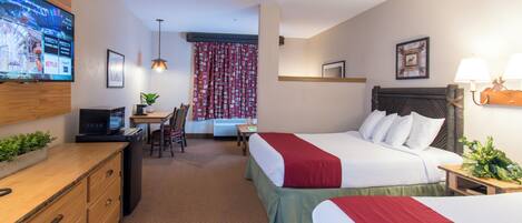 A hotel room with two beds, a desk with a computer, a TV mounted on the wall, and a small dining area.