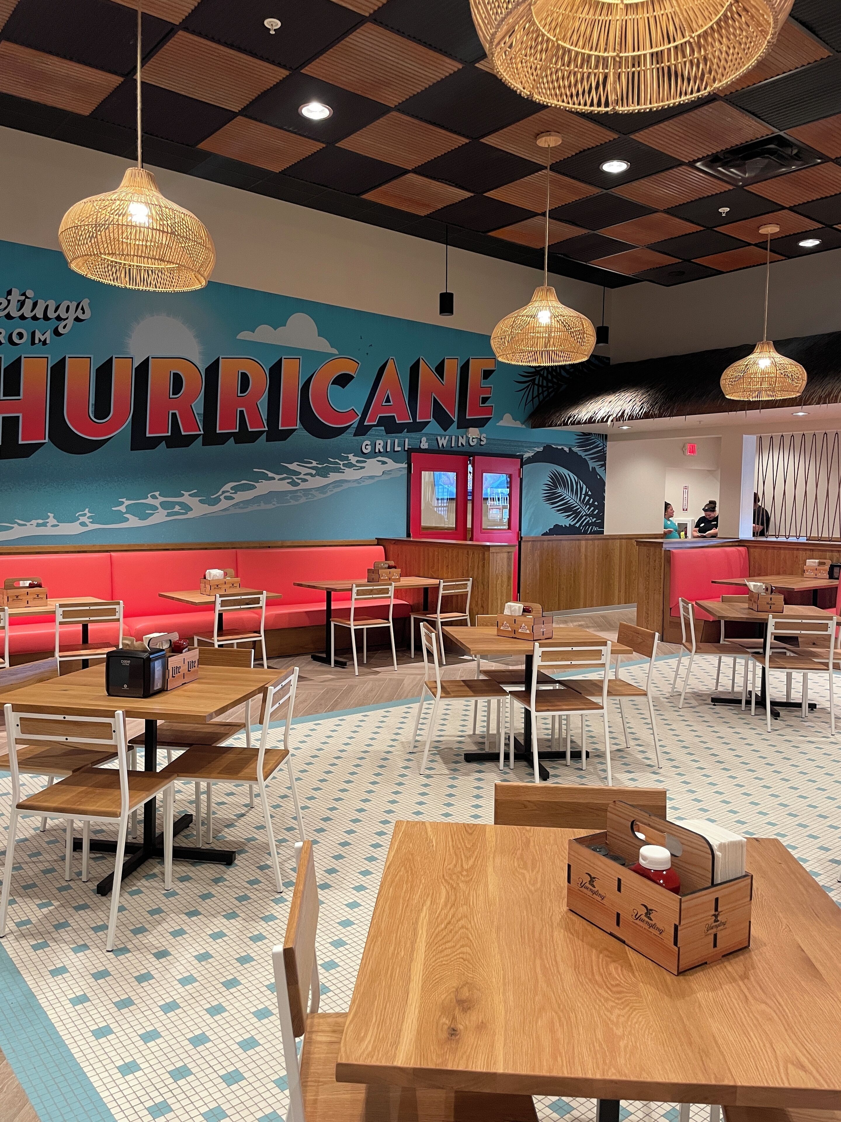 A restaurant interior with wooden tables and chairs, a large mural with the word "HURRICANE," and hanging wicker lamps.