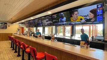 A sports bar with multiple screens displaying sports events, red bar stools, and a counter with a 'Lite' beverage dispenser.