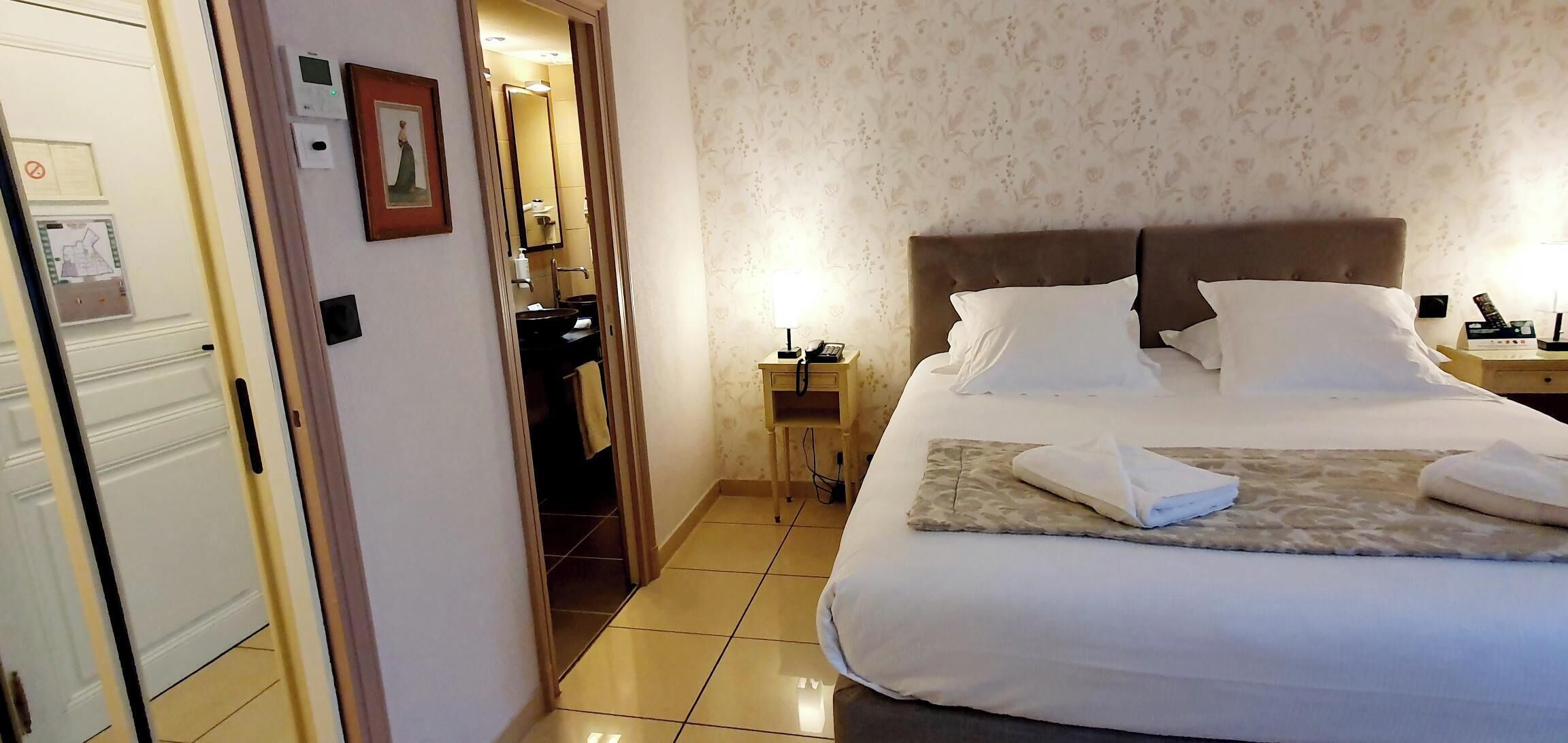 A hotel room with a large bed, a bedside table with a lamp, and a bathroom visible through an open door.