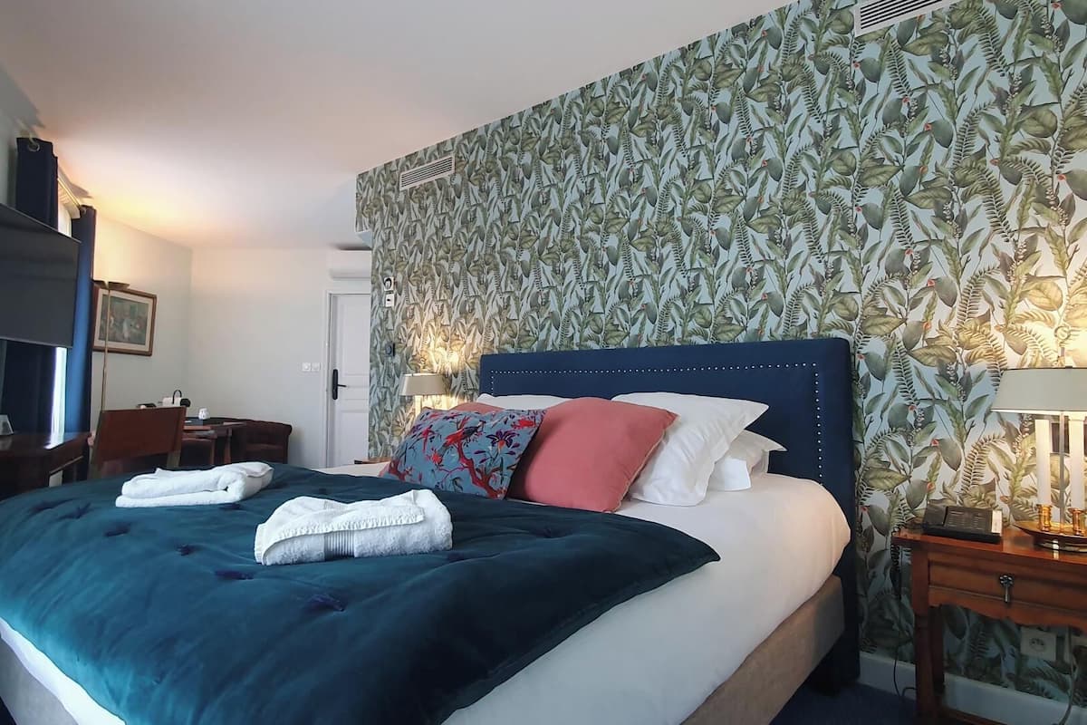 Una cama cuidadosamente arreglada con varias almohadas y una manta azul, una mesita de noche con una lámpara, y una pared con un estampado de hojas.