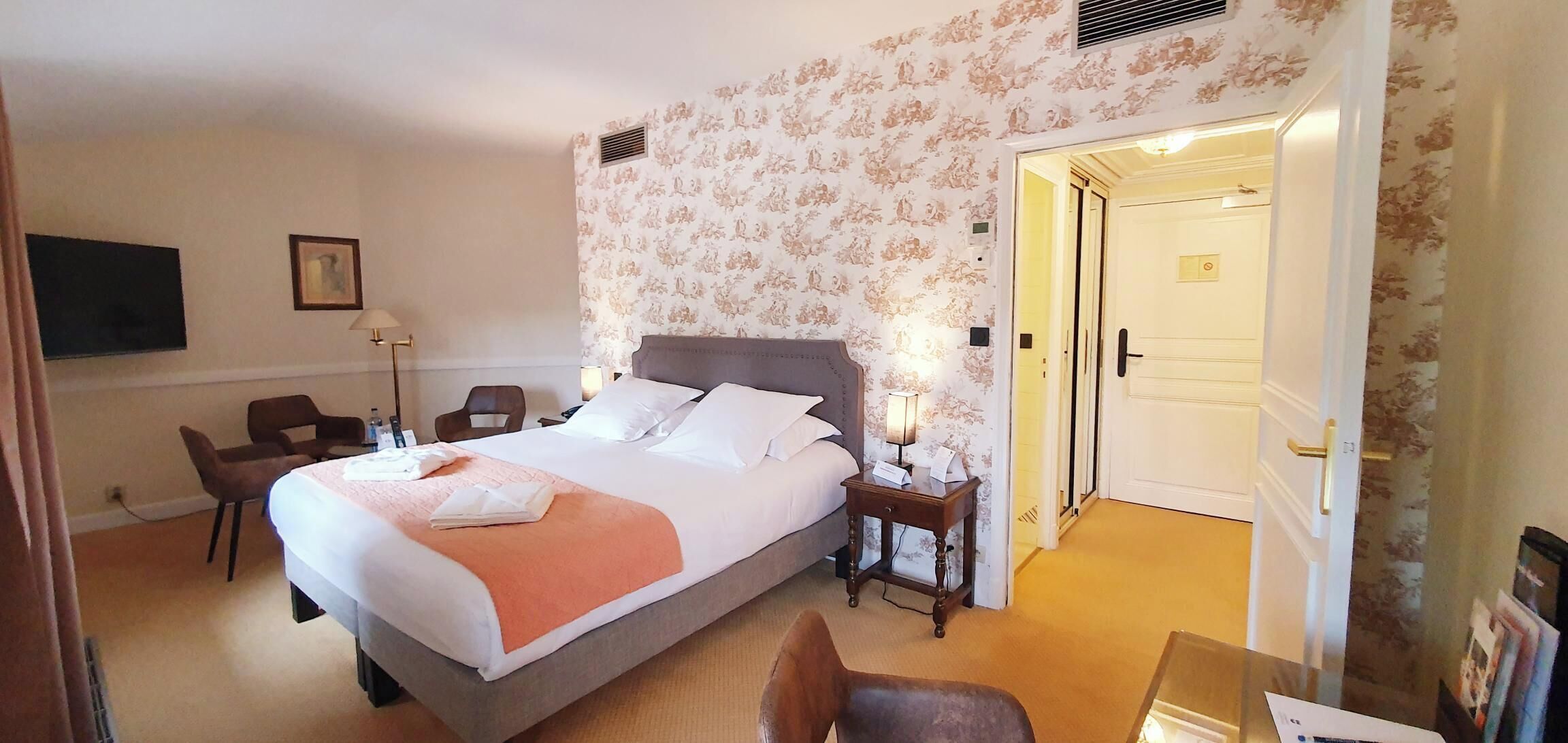 A hotel room with a large bed, a desk with a chair, a small table with a lamp, and a door leading to another room.