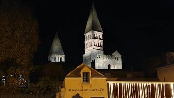 Ein historisches Gebäude mit zwei markanten Türmen, einer mit spitzer und der andere mit runder Kuppel. Im Vordergrund befindet sich ein Gebäude mit einem Schild, auf dem