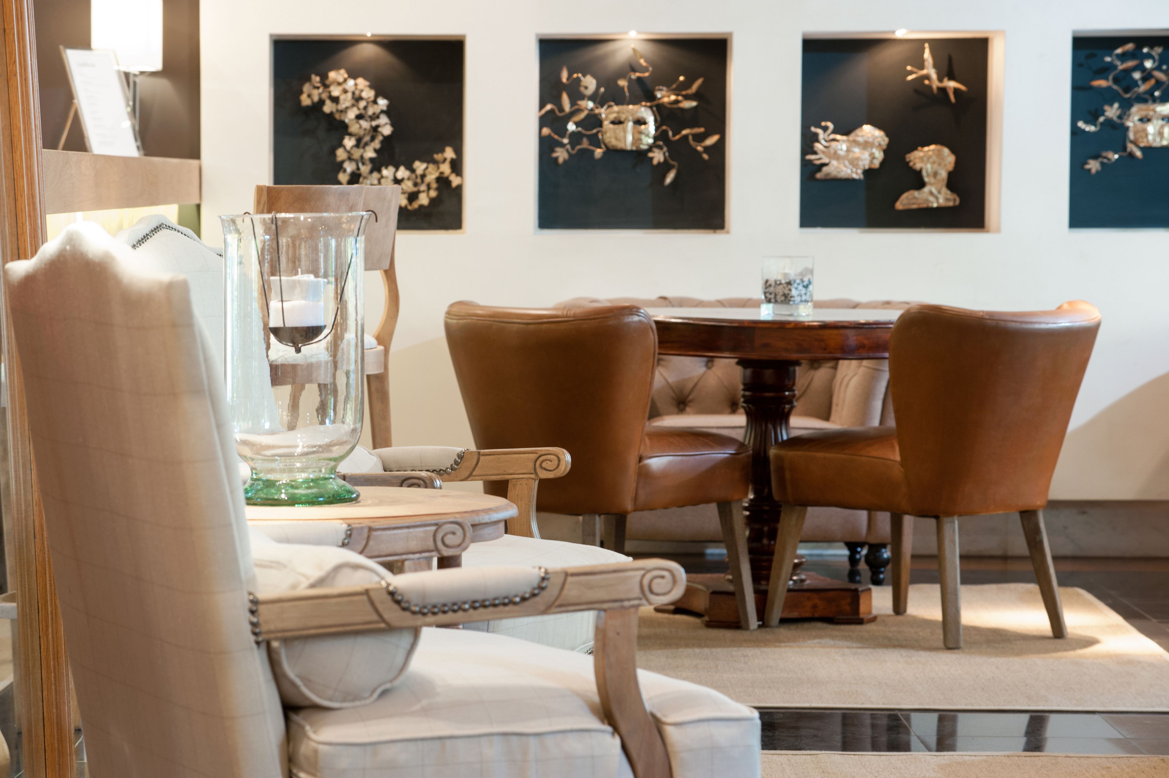 A dining area with a round table and four brown chairs. There are framed artworks on the wall behind the table.