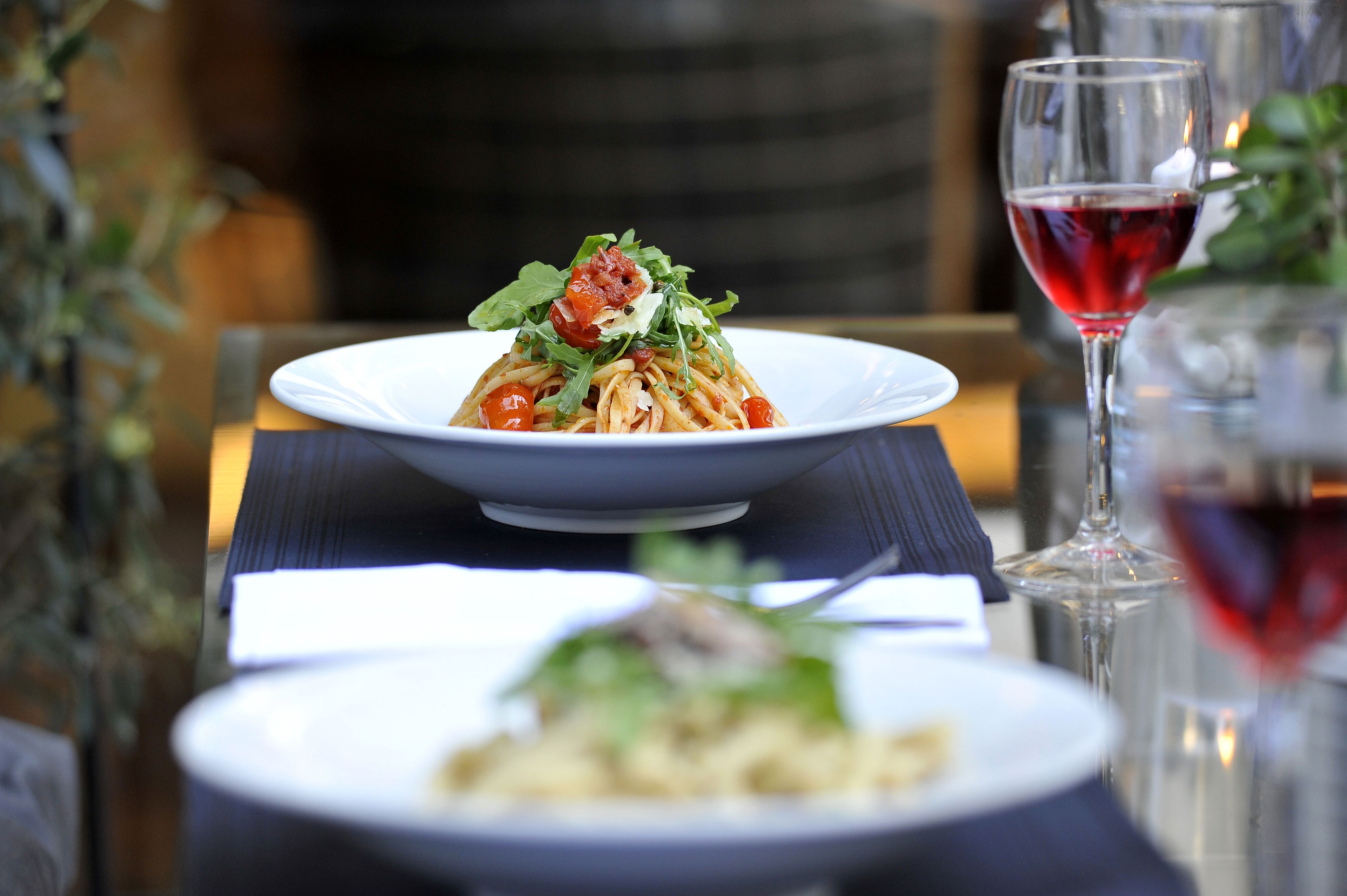 A dining table with a plate of pasta garnished with tomatoes and greens, a glass of red wine, and another plate with a different dish.