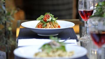 A dining table with a plate of pasta garnished with tomatoes and greens, a glass of red wine, and another plate with a different dish.