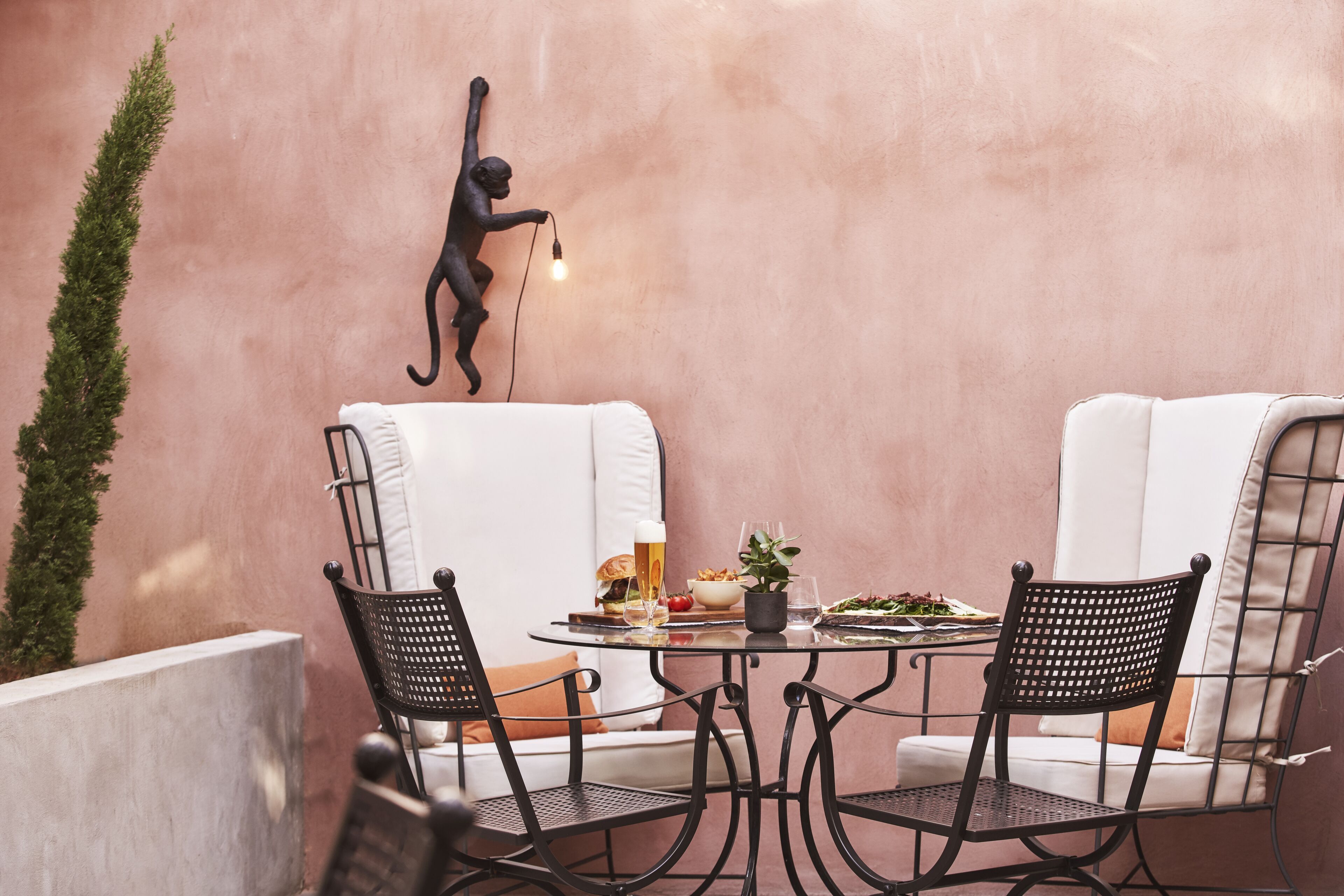 Een terras met een ronde tafel, twee zwarte wickerstoelen, een witte bank met kussens, een wandlamp en een bloempot.