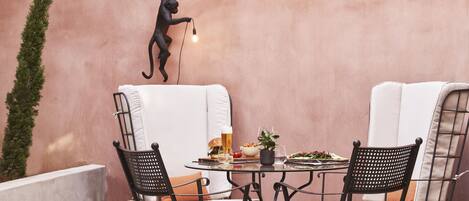 A patio with a round table, two black wicker chairs, a white cushioned bench, a wall-mounted light fixture, and a potted plant.