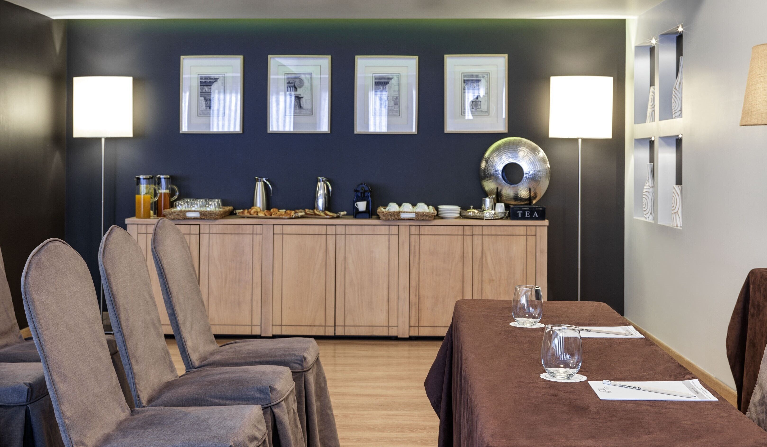 A modern kitchen with wooden cabinets, a countertop with tea and coffee equipment, framed pictures on the wall, and a dining area with a table and chairs.