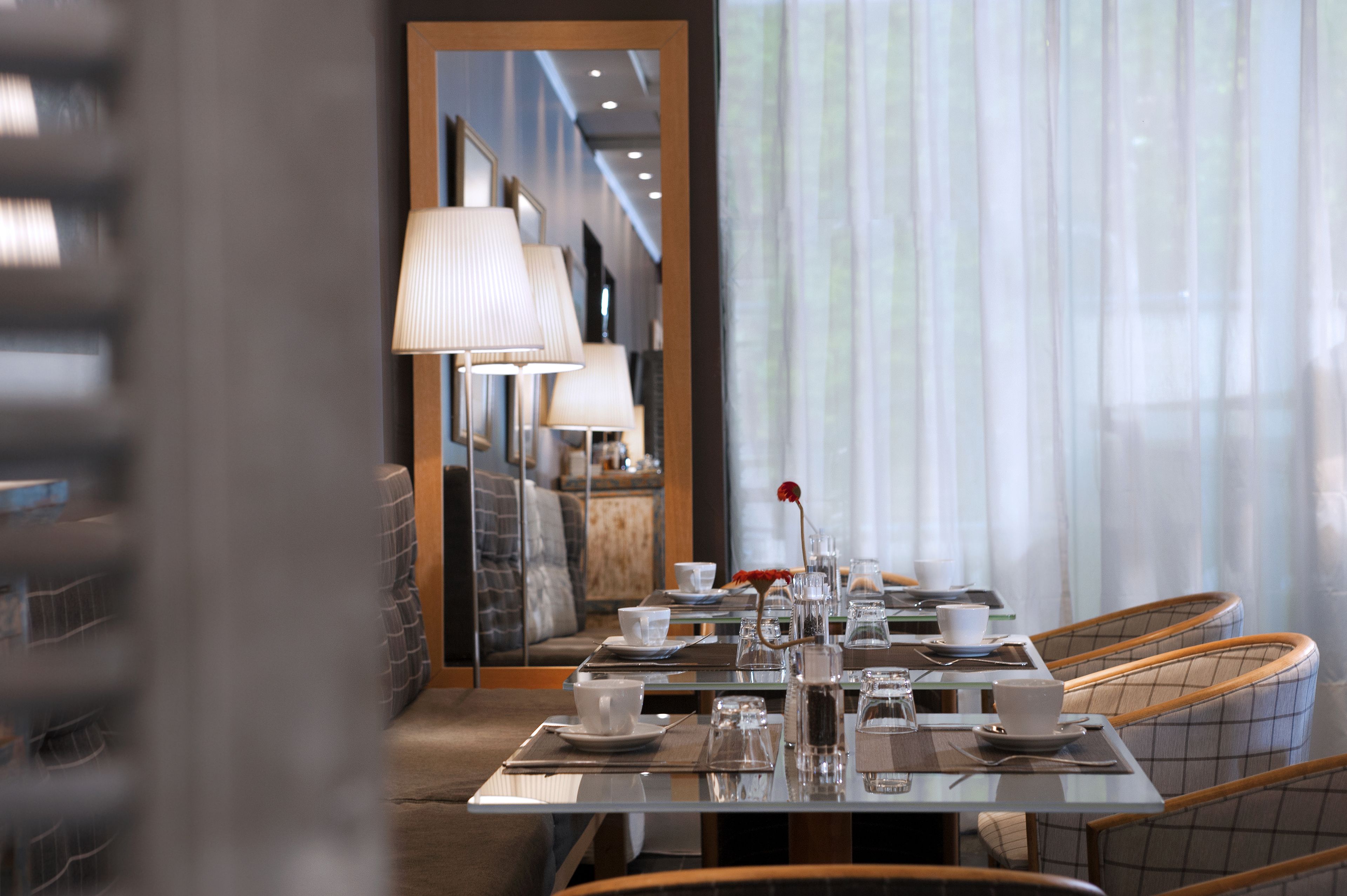 A dining area with a table set for two, featuring a vase with a red flower, a mirror reflecting the room, and a view of the corridor.