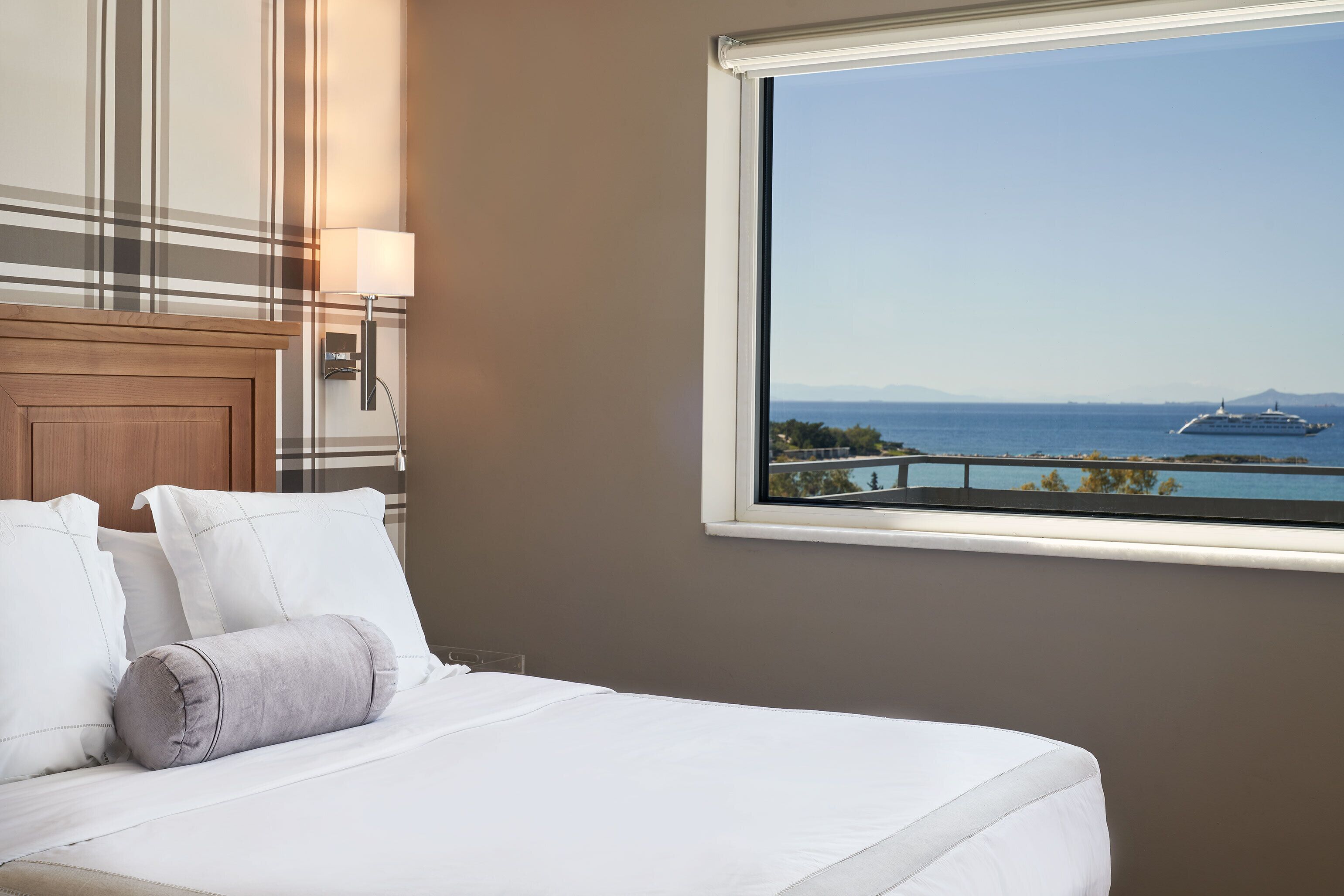 A hotel room with a bed, a window with a view of the sea and a cruise ship, and a wall with a striped pattern.