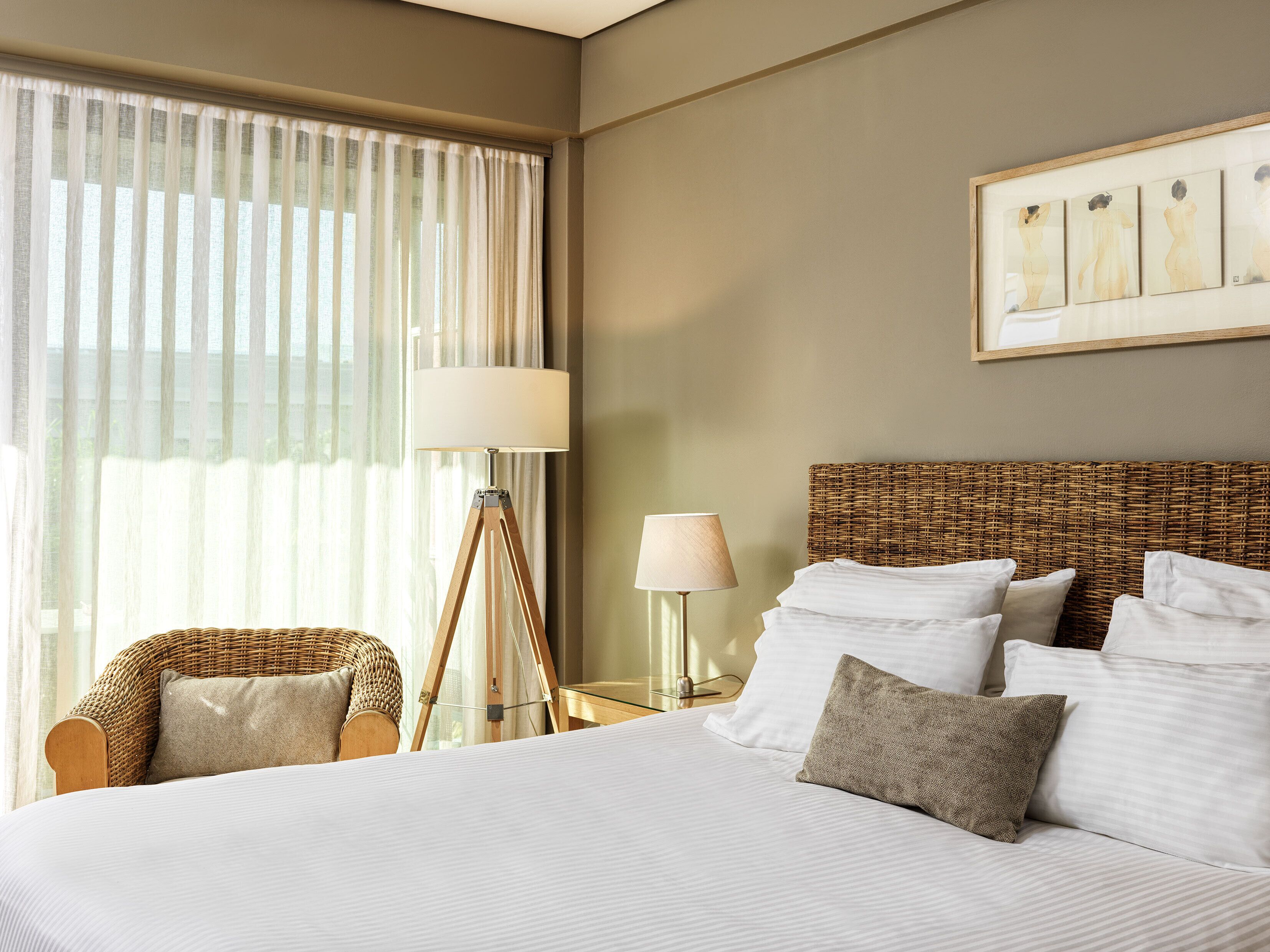 A hotel room with a large bed, a wicker chair, two bedside tables with lamps, and a large window with sheer curtains.