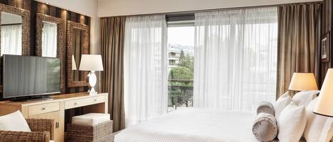 A hotel room with a large bed, a TV, a desk with a lamp, wicker furniture, and a view of the city.