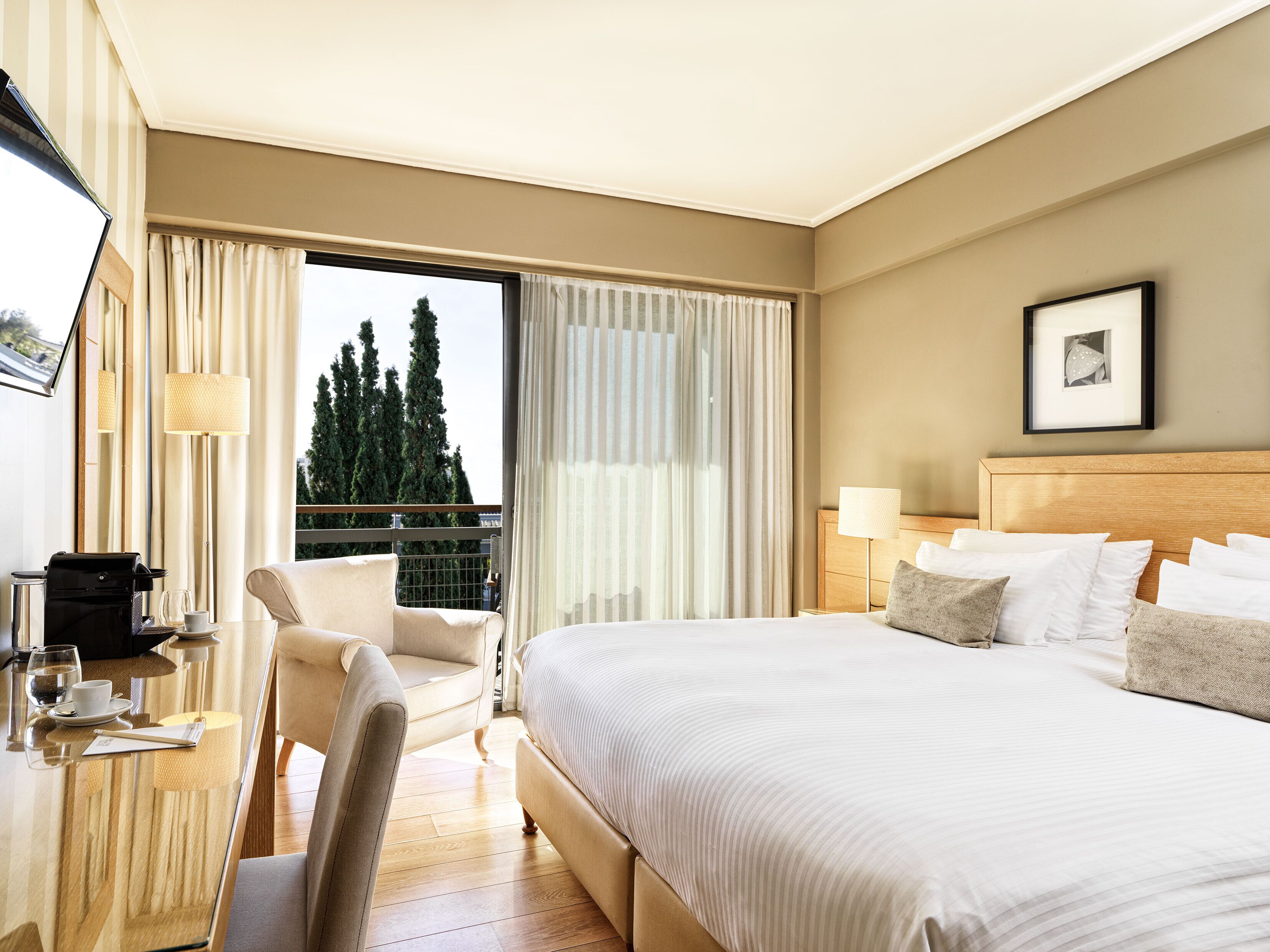 A hotel room with a bed, a desk with a coffee maker, a chair, and a view of trees through the window.