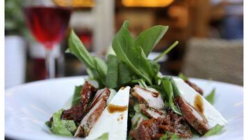 A plate of food with meat, cheese, and greens, with a glass of red wine in the background.