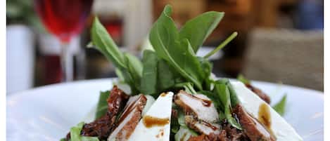 A plate of food with meat, cheese, and greens, with a glass of red wine in the background.