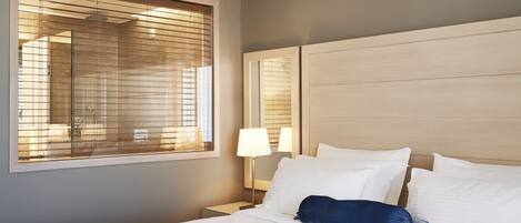 Una habitación de hotel con un ventanal grande, una cama con ropa de cama blanca, una lámpara de noche y un cabecero de madera.