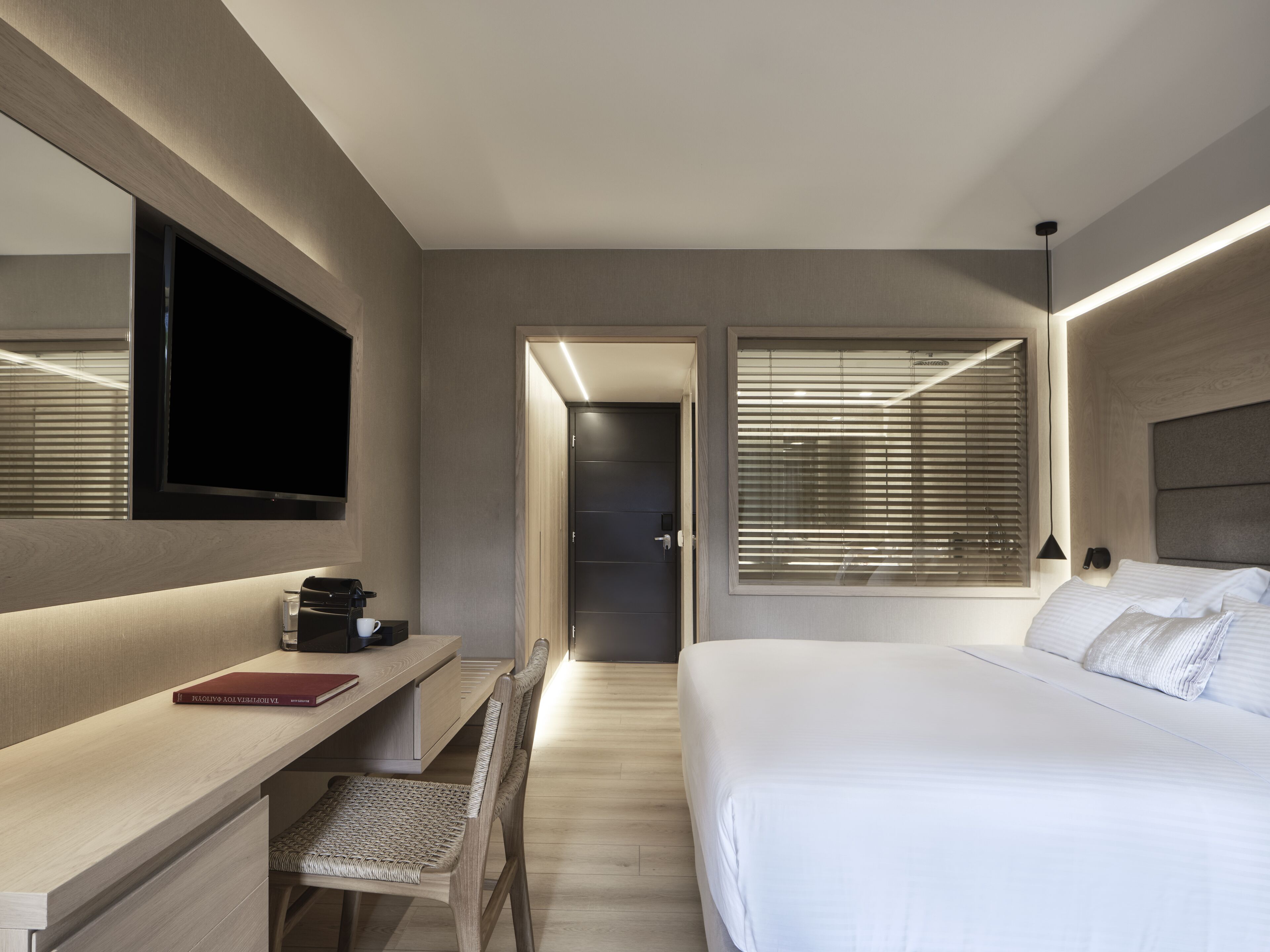 A modern hotel room with a bed, a desk with a chair, a TV, and a window with blinds.