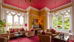 A room with a red carpet, multiple sofas, a piano, and large windows with a garden view. - Cotford Hotel (Malvern)