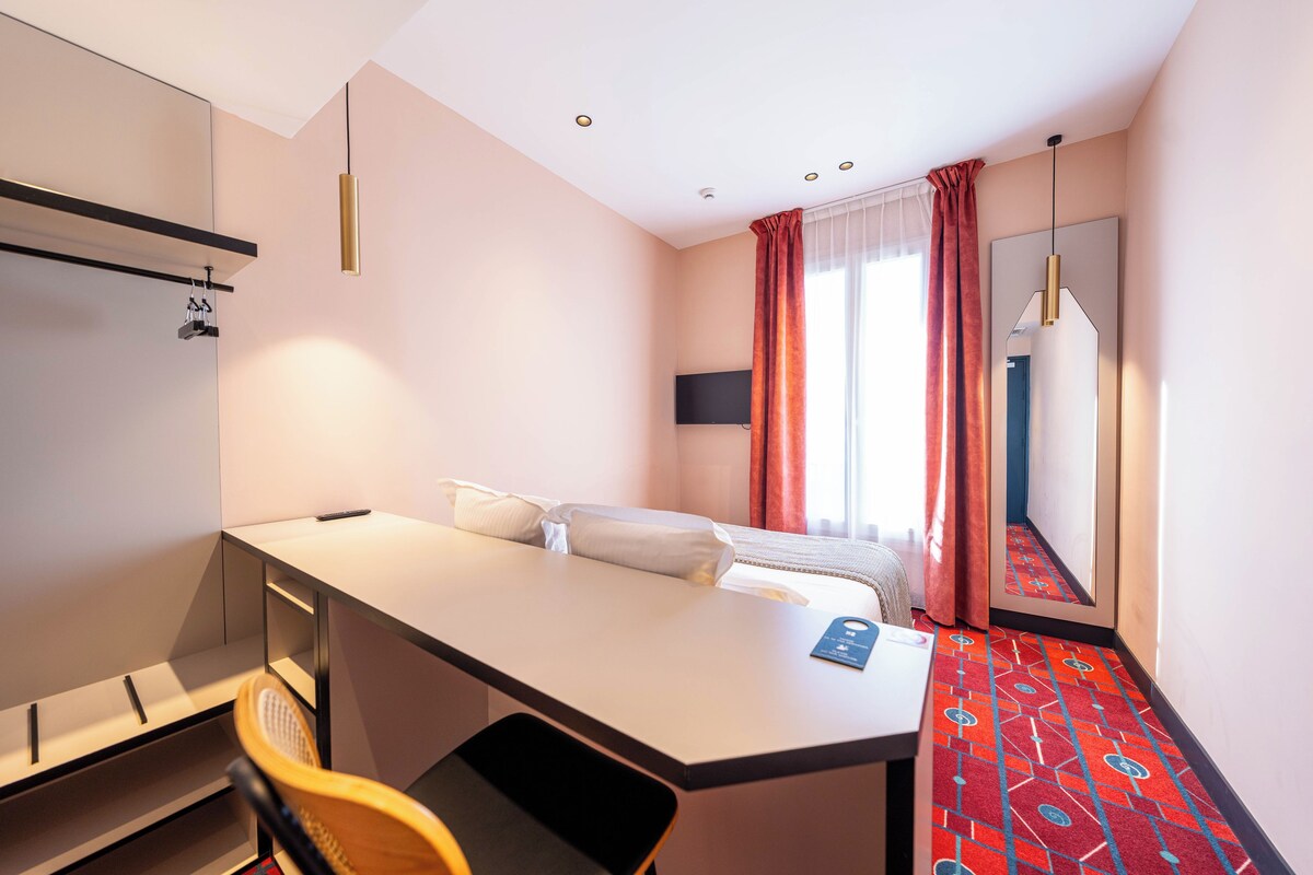 A hotel room with a bed, a desk, a chair, a mirror, and a window with curtains.