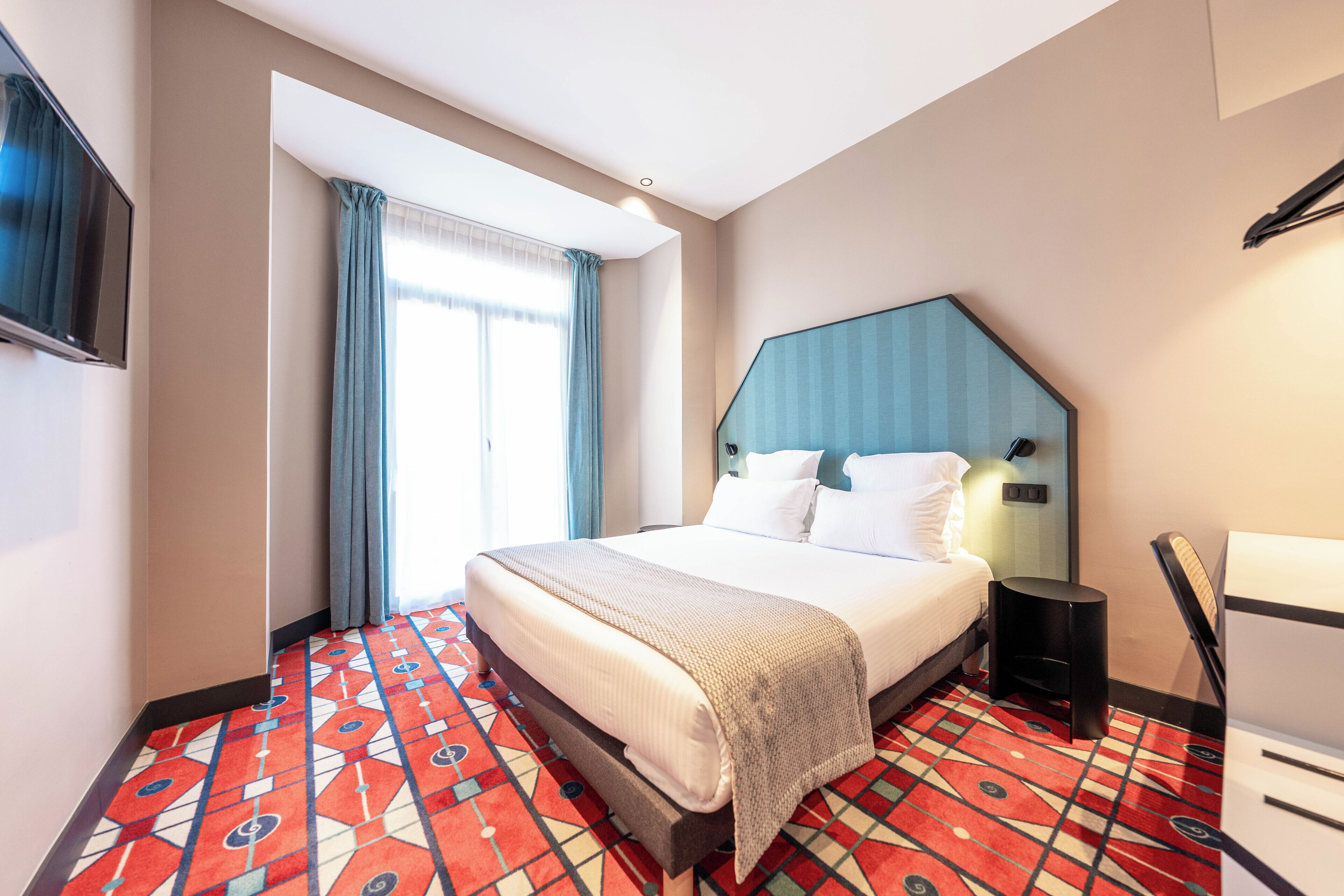 A hotel room with a bed, a TV, a desk, and a window with curtains.