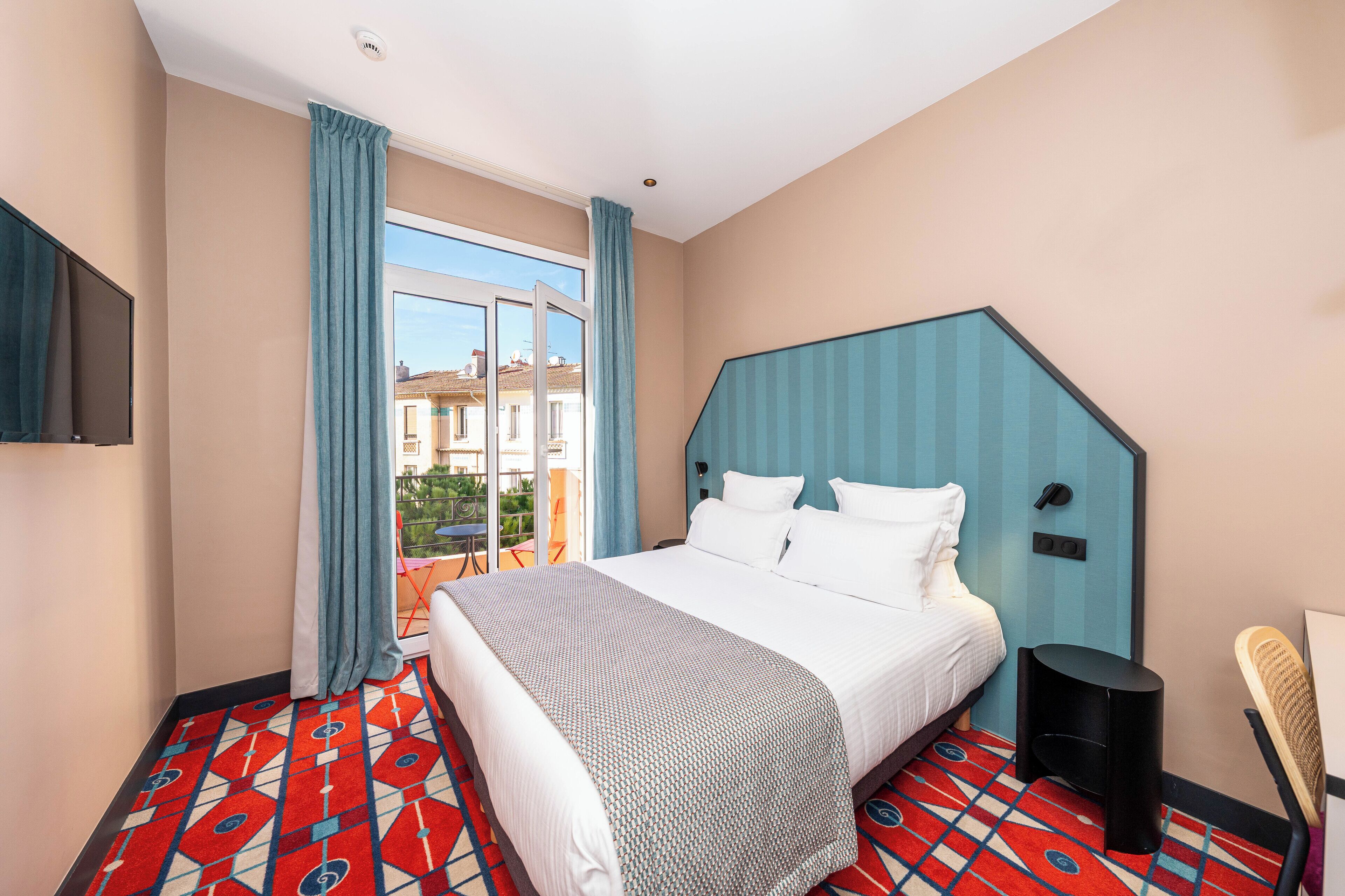 Een hotelkamer met een groot bed, een aan de muur bevestigde televisie, een raam met gordijnen en een kamerbreed tapijt met een patroon.