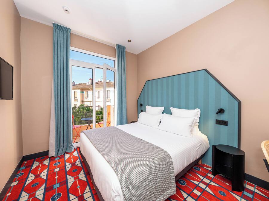 Een hotelkamer met een groot bed, een aan de muur bevestigde televisie, een raam met gordijnen en een kamerbreed tapijt met een patroon.