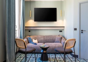 A modern living room with a grey sofa, two wooden chairs, a small coffee table, and a flat-screen TV mounted on the wall.