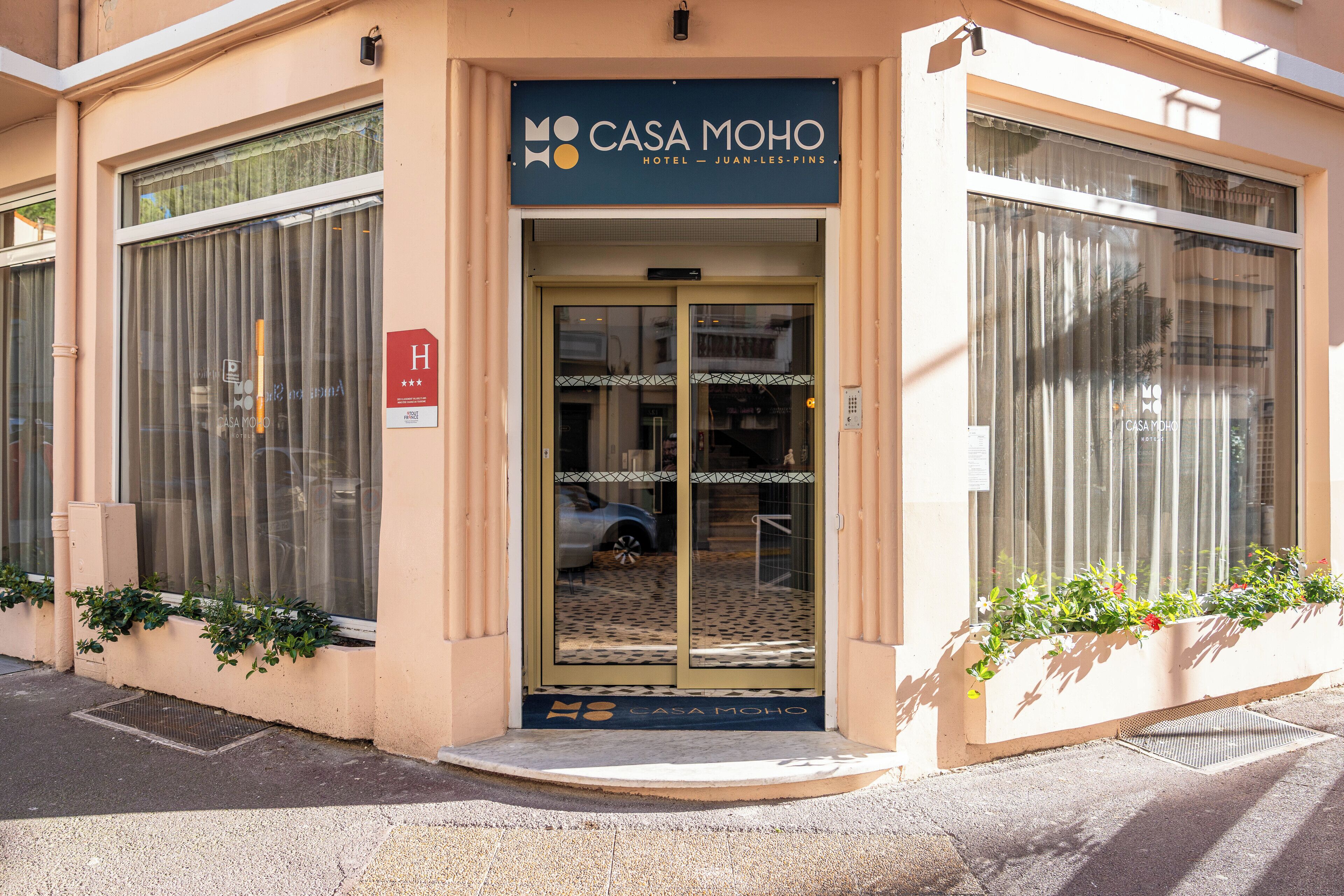 The entrance of Casa Moho Hotel with a glass door, a parking sign, and a small garden area with plants.