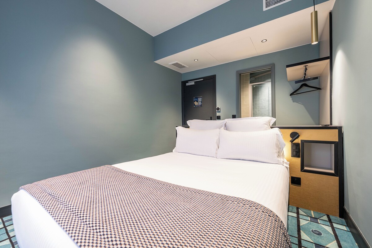 A hotel room with a bed, a bedside table, a mirror, and a hanging coat rack.