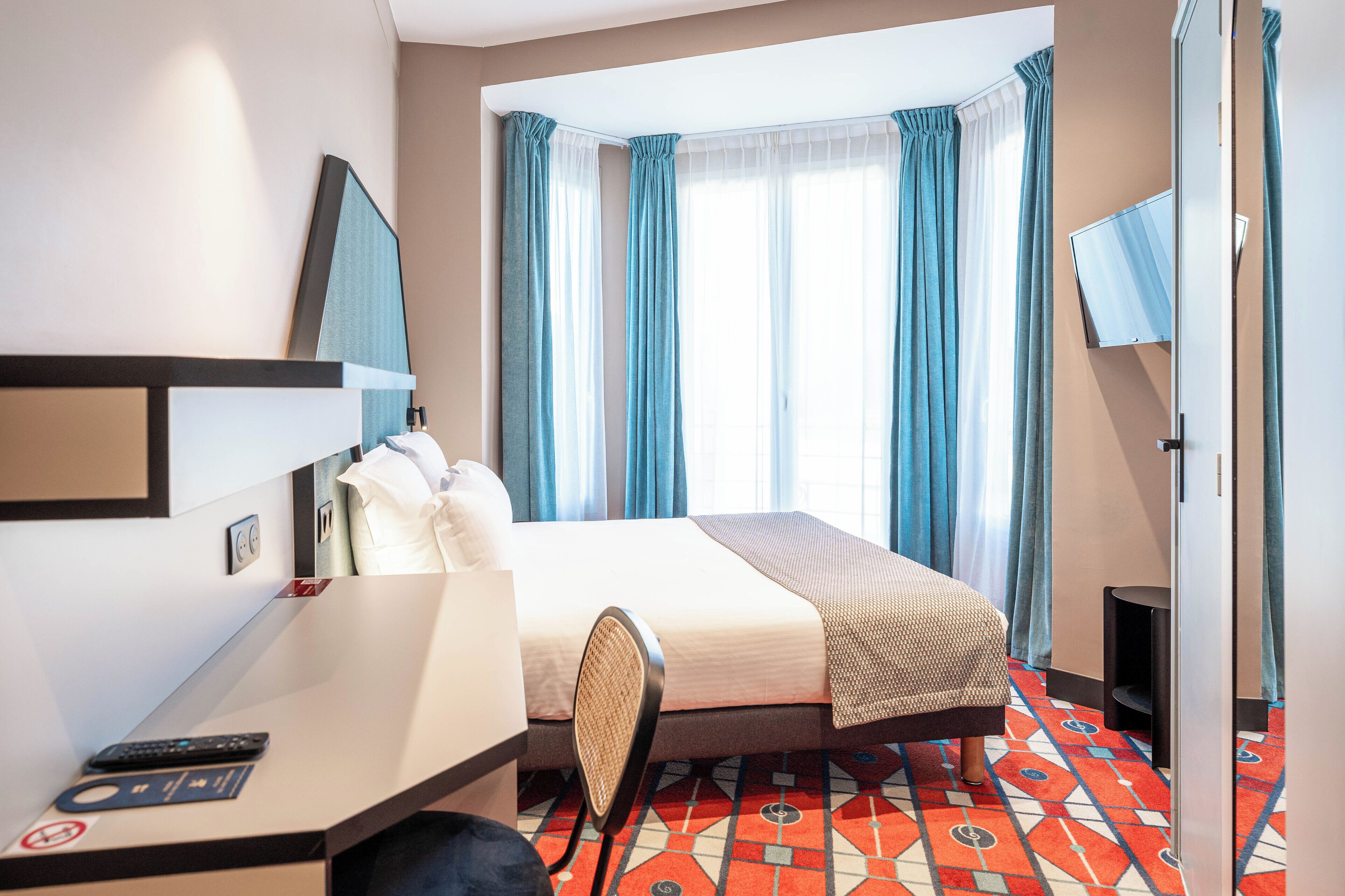 A hotel room with a bed, a desk with a chair, a TV, and a window with curtains.