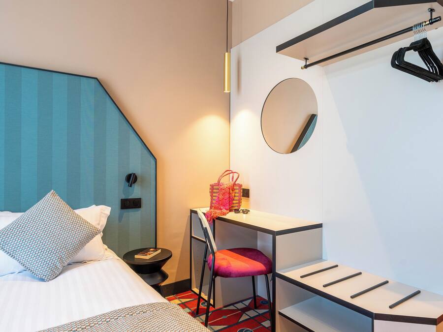 Een hotelkamer met een bed, een bureau, een stoel, een klein tafeltje, een spiegel en een kapstok.