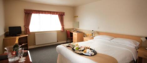 A hotel room with a bed, bedside table, a desk with a chair, a television, a window with curtains, and a radiator.