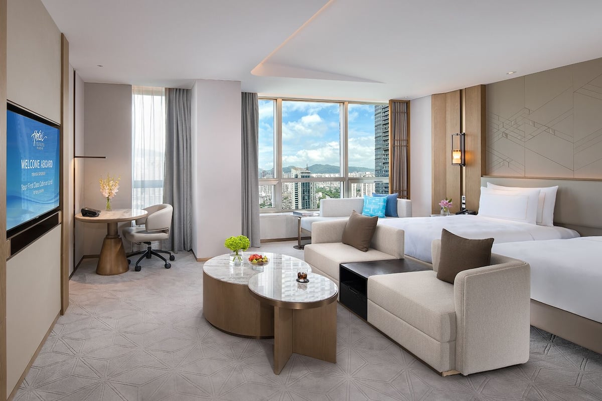 Une chambre d’hôtel moderne, équipée d’un lit, d’un canapé, d’un fauteuil et d’une petite table sur laquelle se trouvent un vase et des fruits.