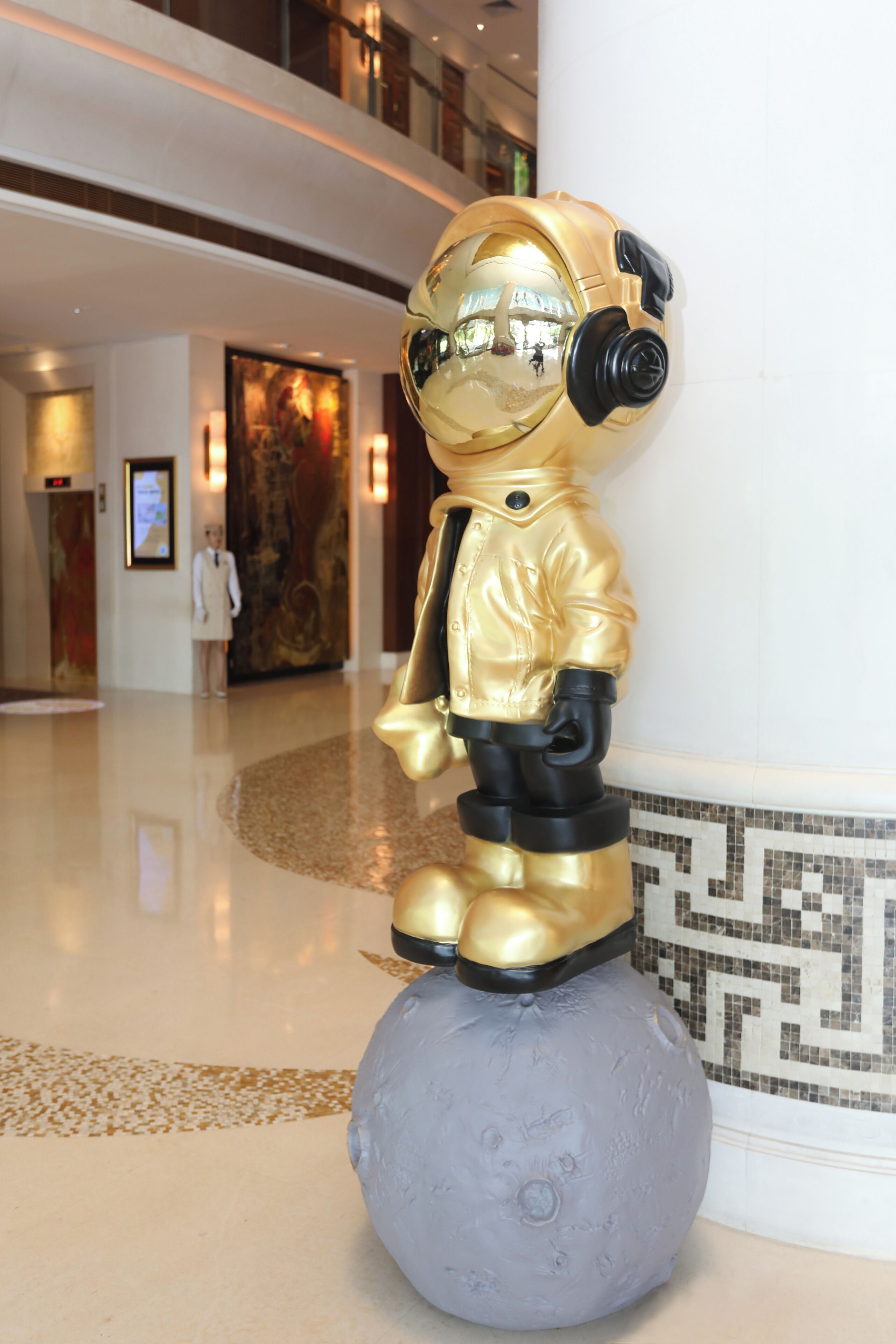 A golden statue of a figure with a helmet and headphones, standing on a spherical base, in a lobby with a tiled floor and a large painting in the background.