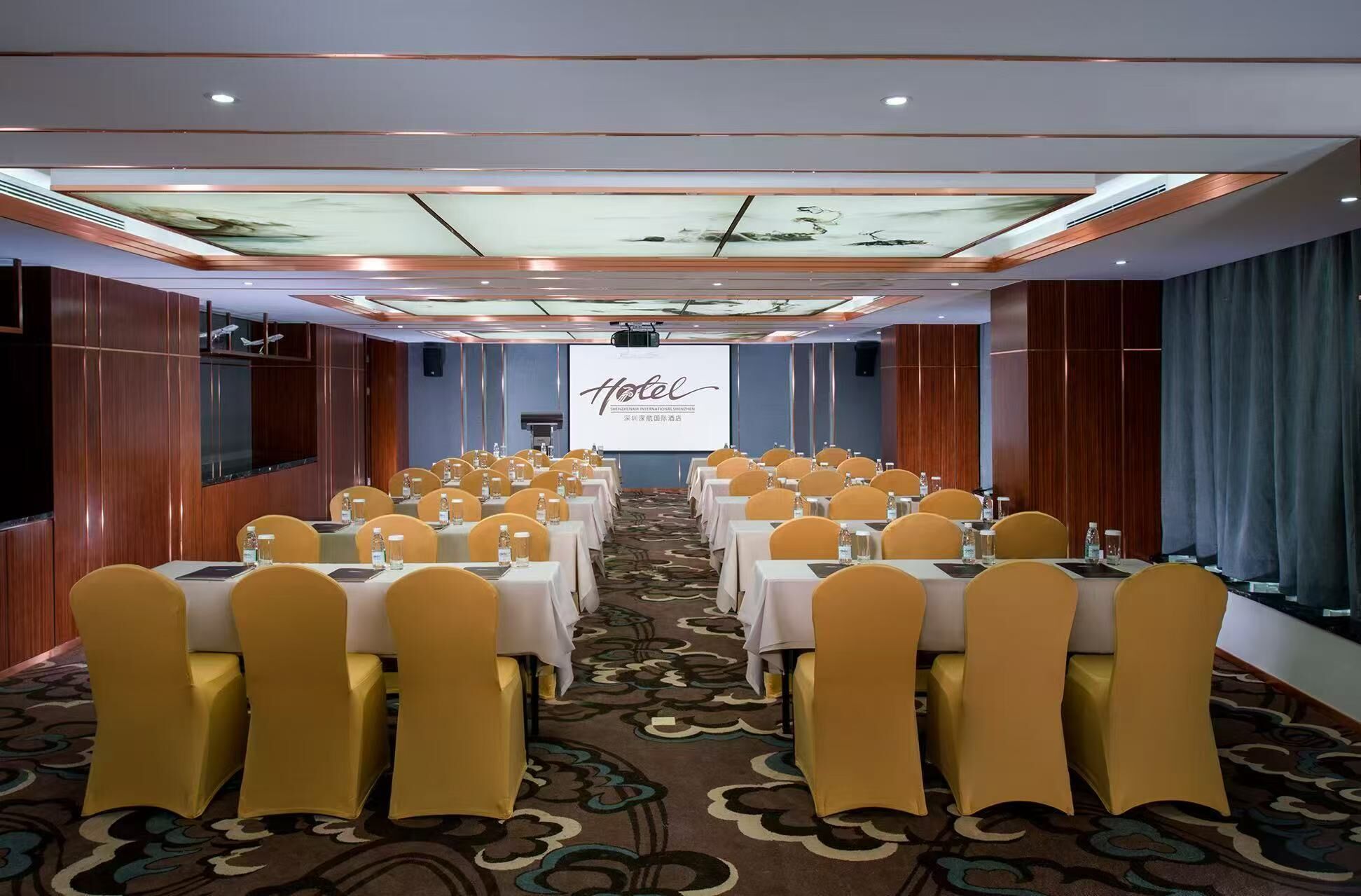A conference room with rows of tables and chairs, a projector screen, and a carpeted floor.