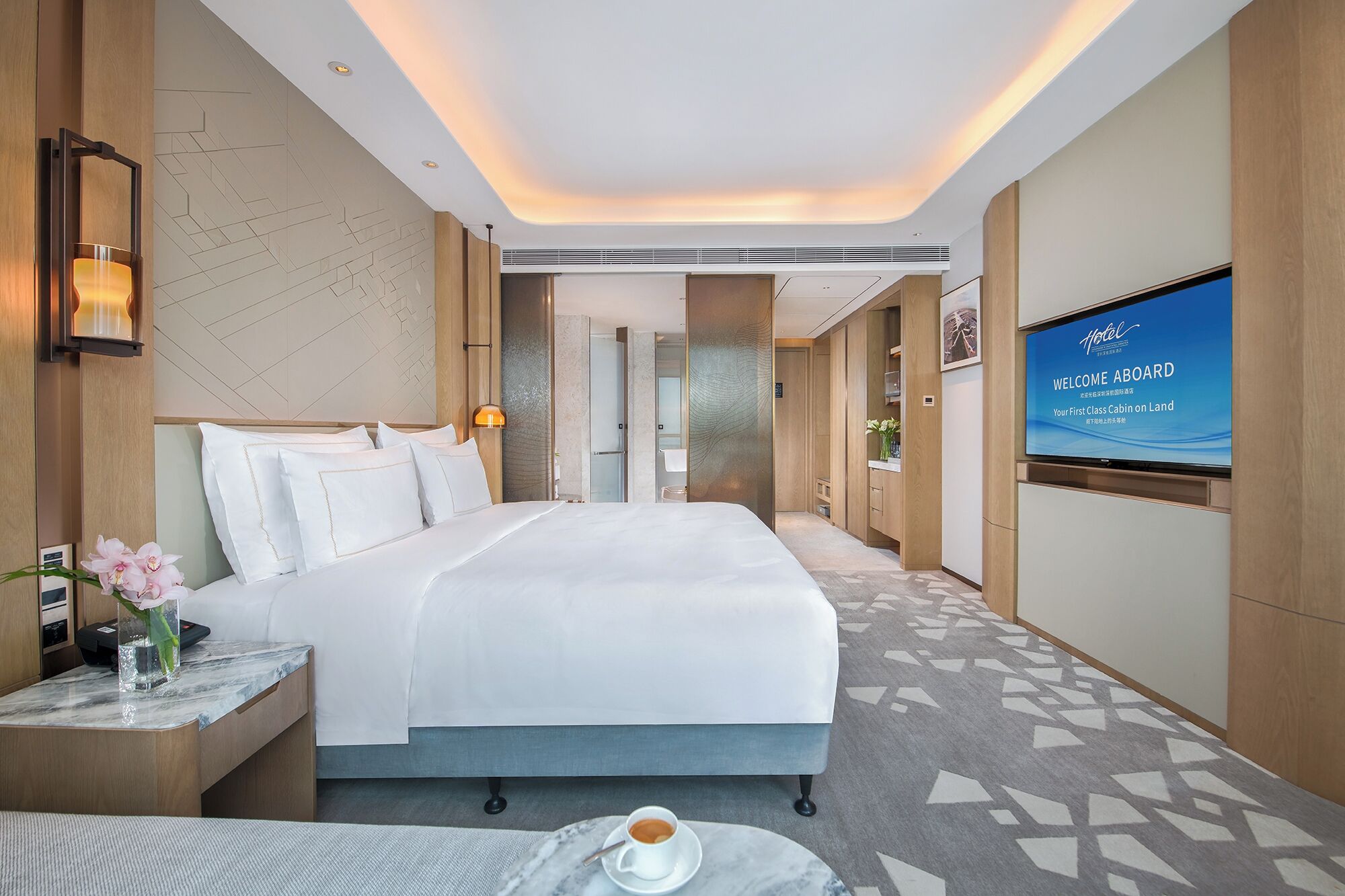 A modern hotel room with a large bed, a television displaying a welcome message, and a view of the corridor.