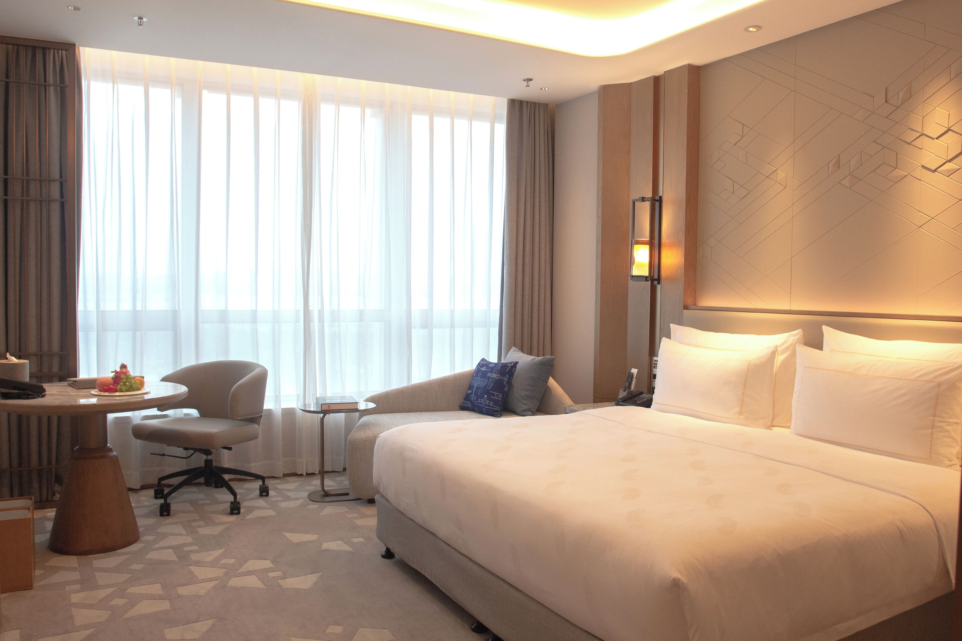 A hotel room with a large bed, a desk with a chair, a small table, and a window with curtains.