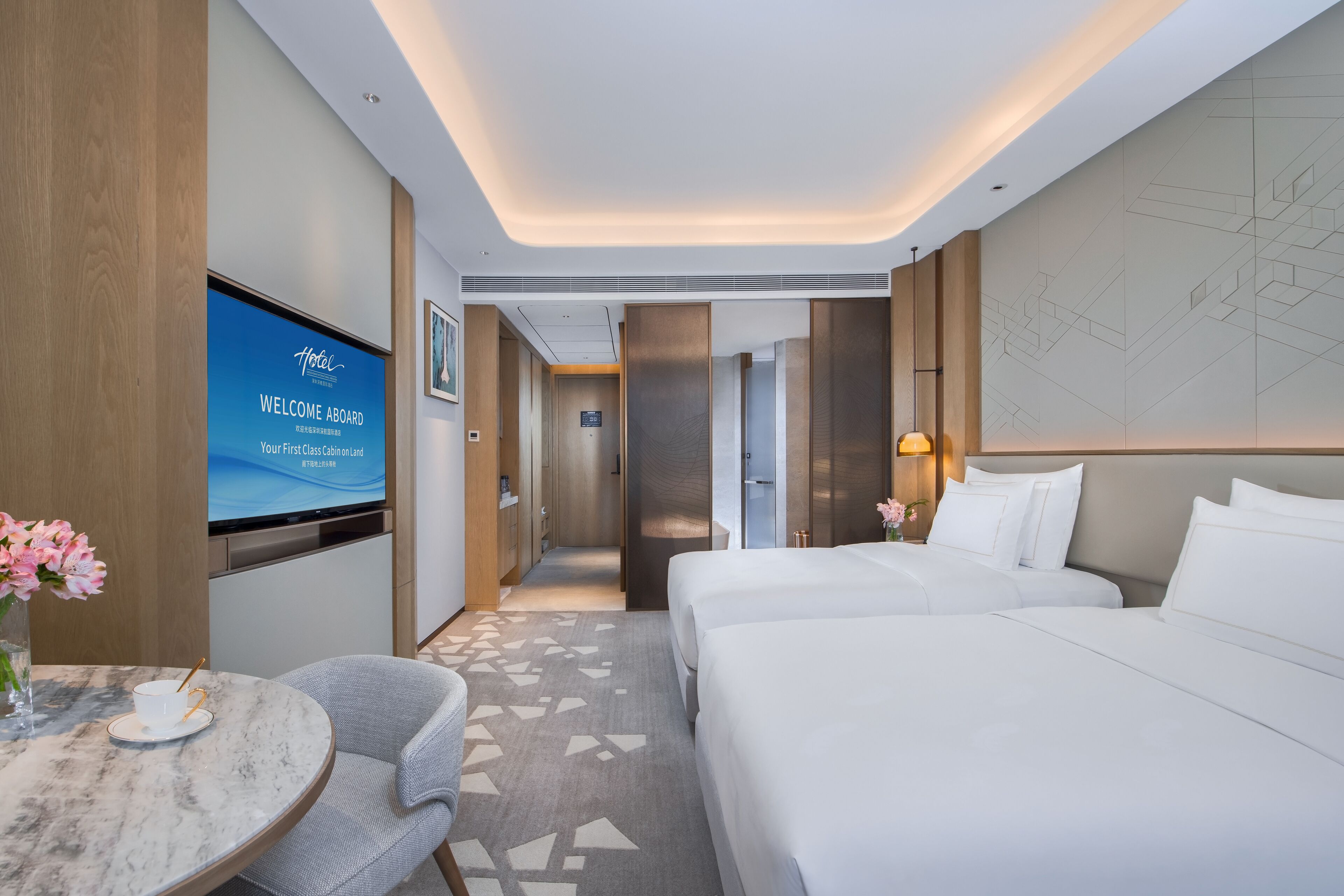 Una habitación de hotel moderna con una cama grande, un pequeño espacio para sentarse, un televisor mostrando un mensaje de bienvenida y un mapa en la pared.