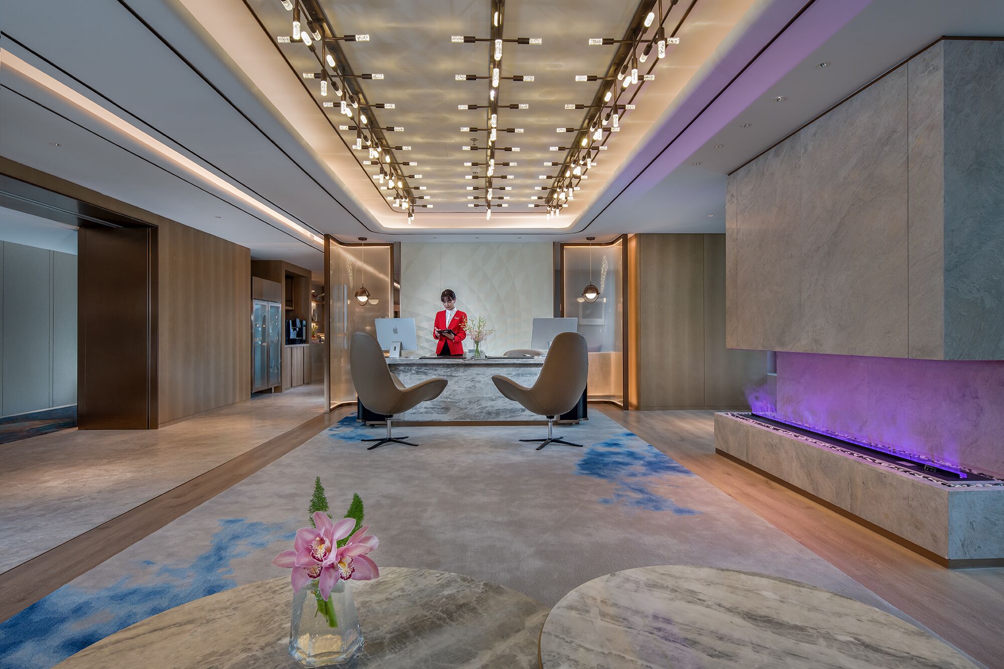 A modern lobby with a reception desk, two chairs, a vase with flowers, and a purple-lit water feature.